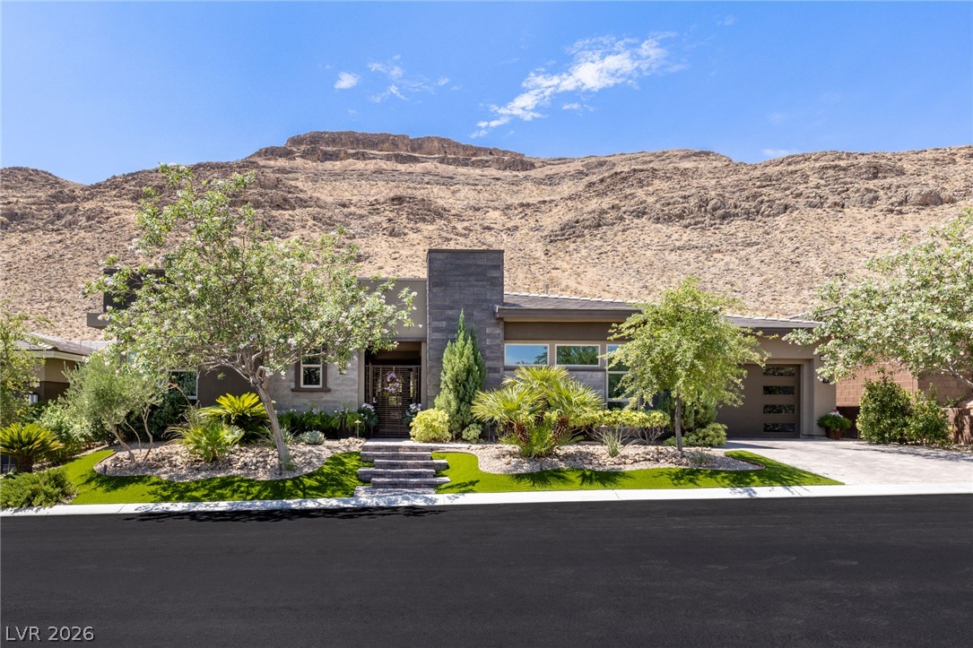 10167 Copper Edge Road Las Vegas, NV 89148 - Photo 1 of 1 Modern desert architecture with a stone-clad entry, framed by mature trees and a manicured landscape, rests against a majestic mountain backdrop.
