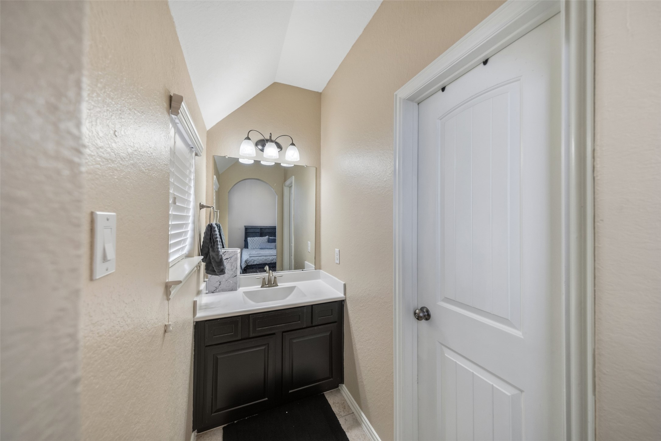 4747 Abercorn Street Rosharon, TX 77583 - Photo 28 of 46 This photo showcases a compact bathroom with a modern design. It features a sleek dark vanity with a white countertop and a large mirror with overhead lighting. The space is well-lit with natural light from a window, and there's a white door leading to an adjacent area.