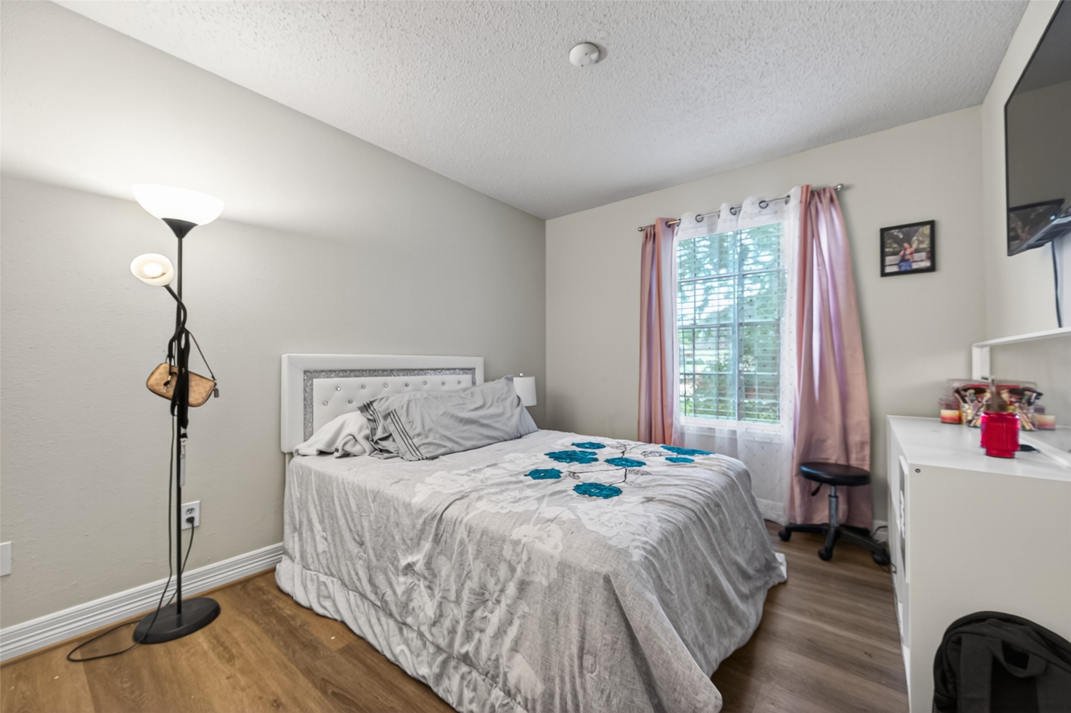 18446 North Willow Bluff Road Katy, TX 77449 - Photo 27 of 33 This cozy bedroom features a comfortable bed with a tufted headboard, soft lighting, and wood flooring. A large window with pink curtains allows natural light to fill the room. A small desk area and wall-mounted TV add functionality.