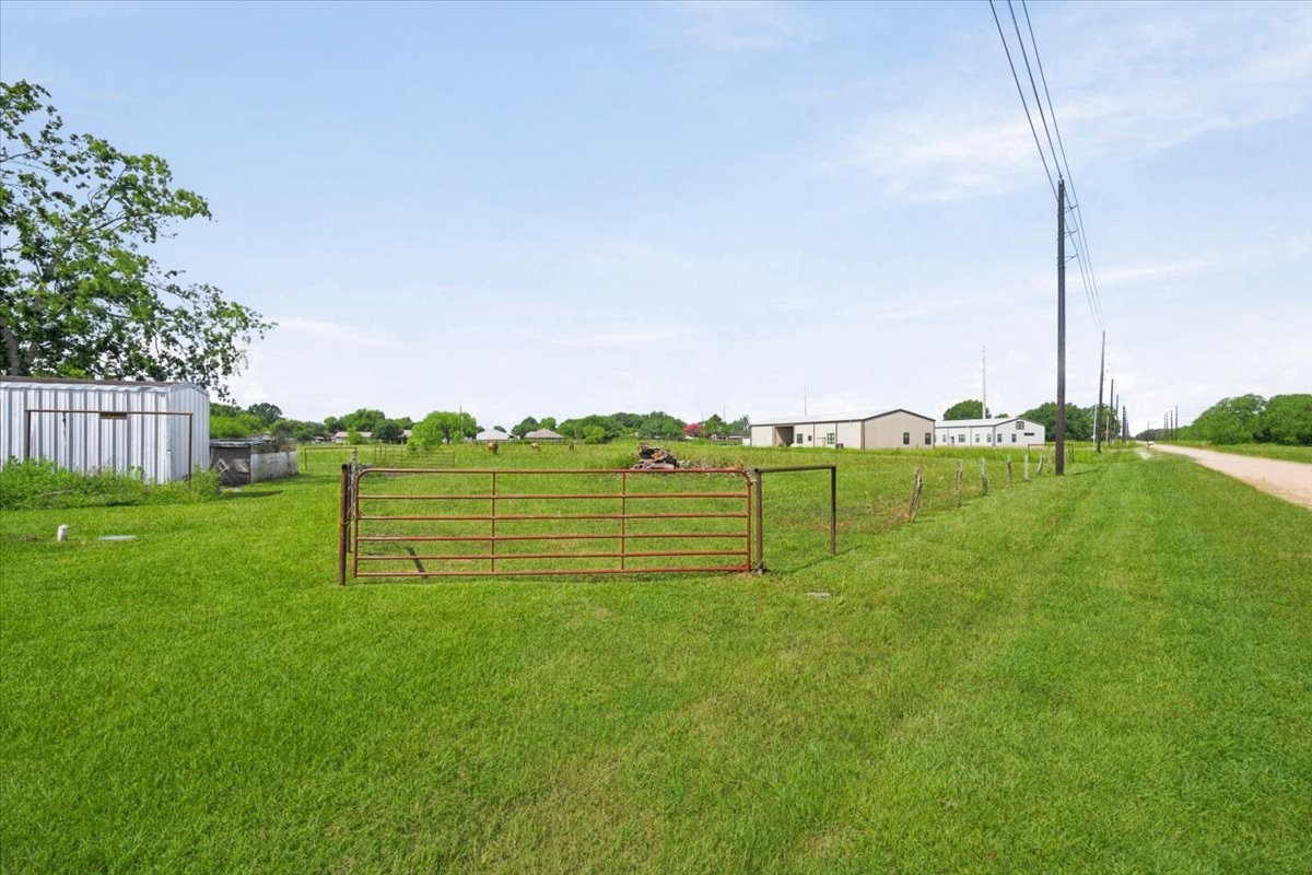 765 Norcross Road Wallis, TX 77485 - Photo 37 of 42 Expansivefield with a rusty gate and fenced boundary, adjacent to a gravel road. Nearby structures include a white building Ideal for those seeking a rural setting with open space.