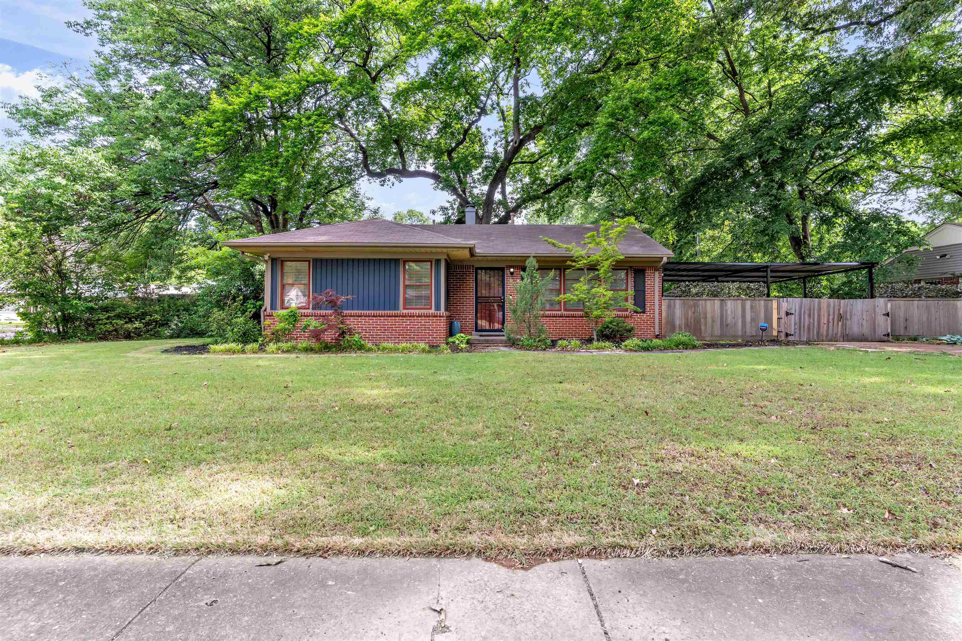 4564 Verne Road Memphis, TN 38117 - Photo 3 of 25 Brick facade residence featuring a front lawn, mature trees, a blue siding accent, an attached carport, and a wooden privacy fence