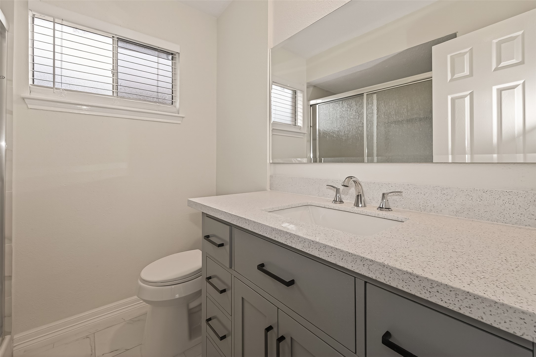 7027 Dickson Way Missouri City, TX 77489 - Photo 20 of 22 This bathroom features a modern design with a light-colored countertop and sleek fixtures. It includes a large mirror, ample cabinet storage, and a window for natural light. The shower has a frosted glass door for privacy.