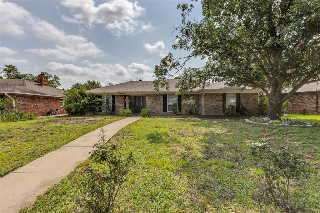 Brick exterior ranch-style home featuring a prominent mature tree, a concrete walkway, and dark window shutters