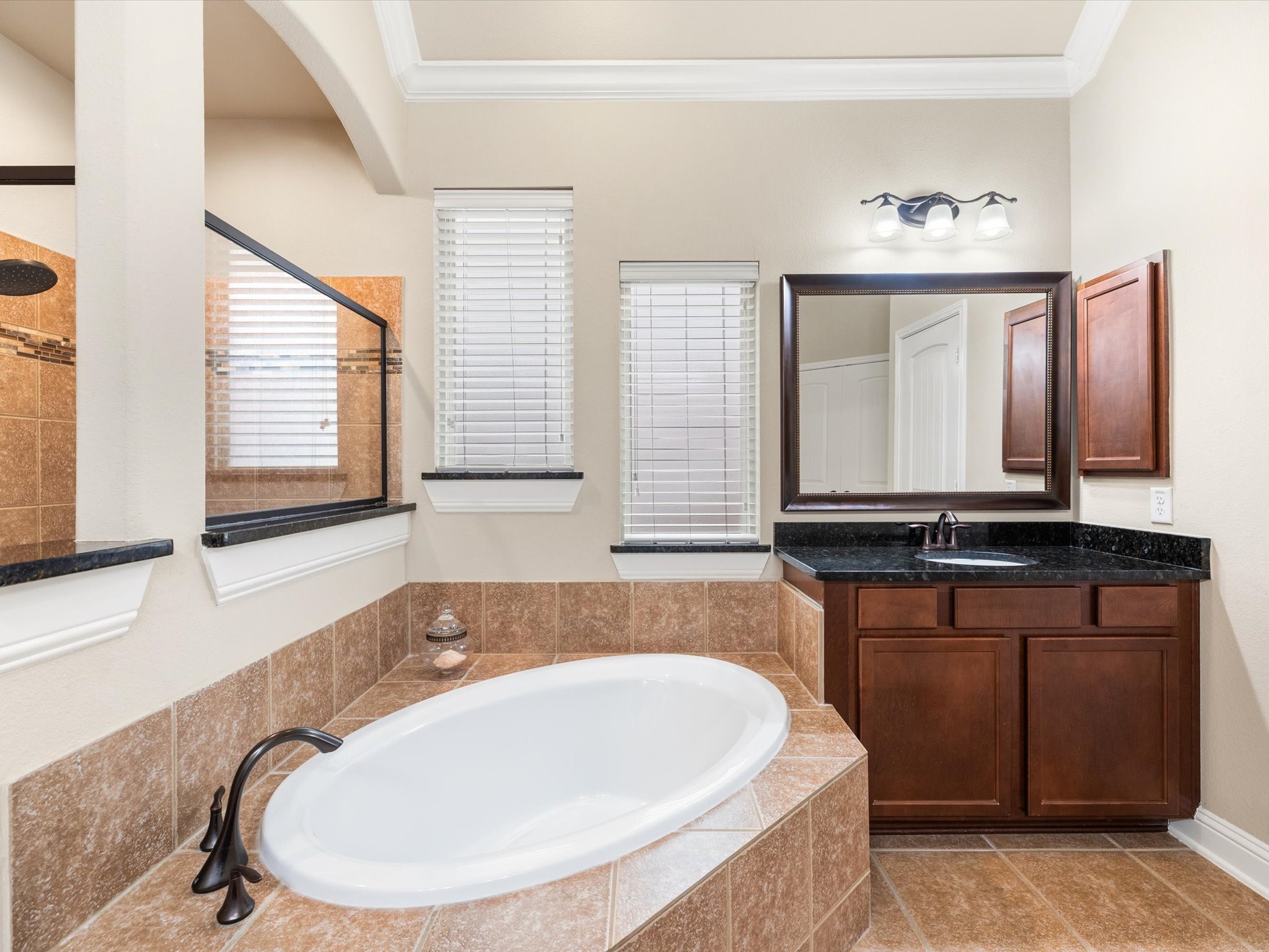 3907 Randle Ridge Court Fulshear, TX 77441 - Photo 20 of 46 This bathroom features a spacious, soaking bathtub with elegant tile surround, a large vanity with dark granite countertops, and rich wood cabinetry. Natural light streams in through two windows, creating a bright and inviting space.