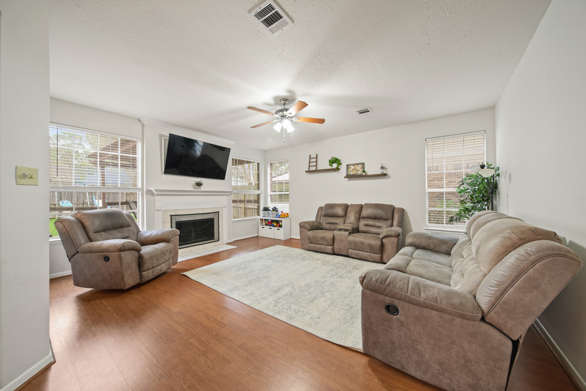16114 Kintyre Point Road Houston, TX 77095 - Photo 16 of 44 Bright and inviting living room with large windows, hardwood floors, and a cozy fireplace. Features include a mounted TV, ceiling fan, and comfortable seating, creating a perfect space for relaxation and entertainment.