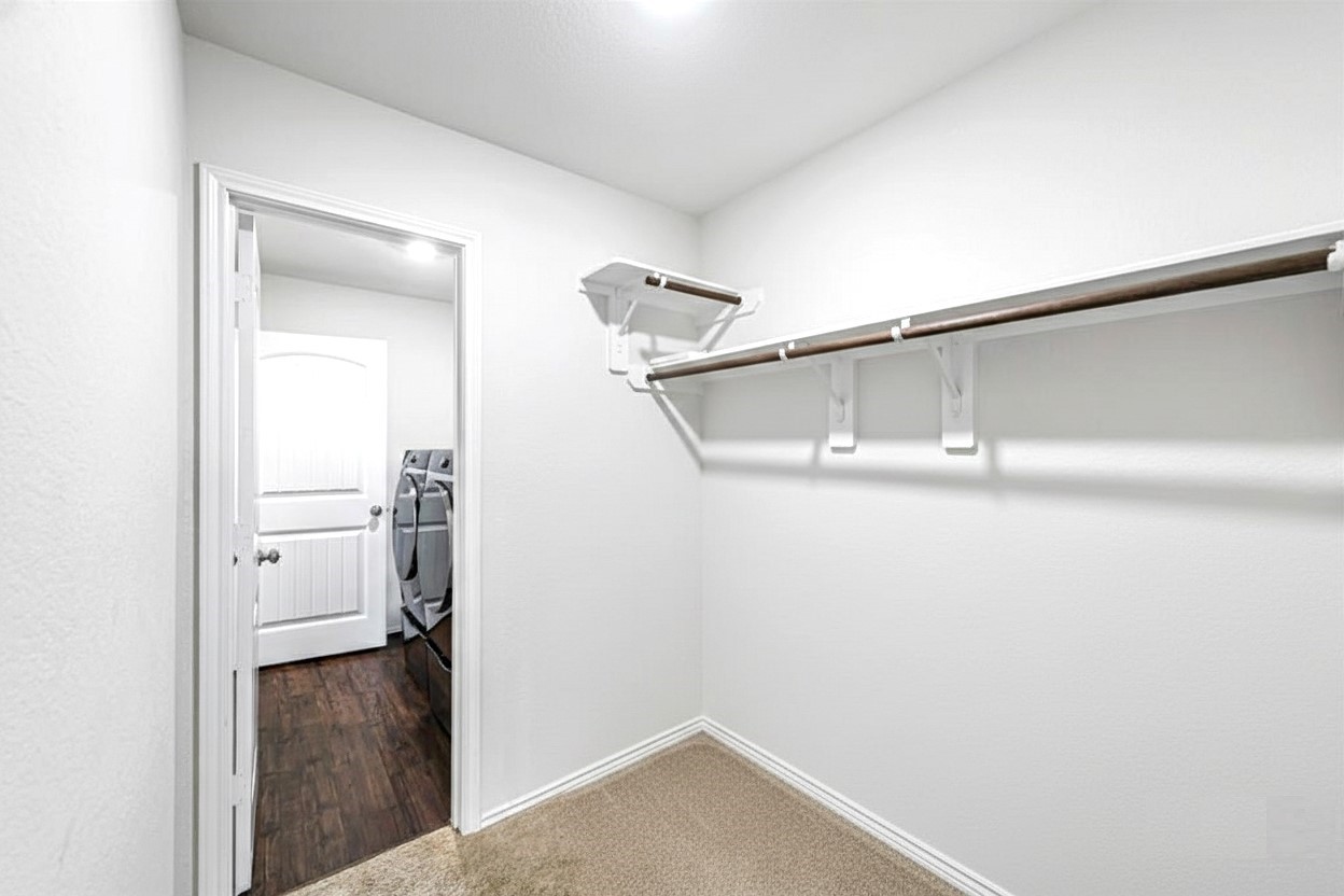 17439 Chester Valley Trail Hockley, TX 77447 - Photo 14 of 22 This photo showcases a spacious walk-in closet with a single hanging rod and shelf, adjacent to a room with dark wood flooring. The closet features neutral walls and carpeting, providing a clean, bright space for storage.