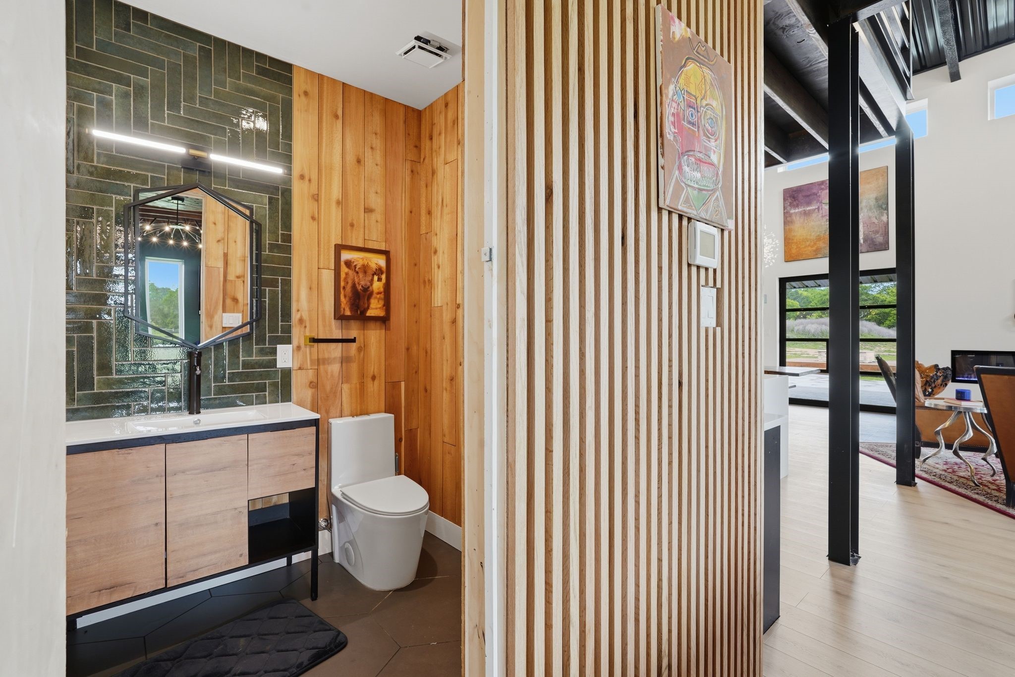 2743 Bobville Road Montgomery, TX 77316 - Photo 44 of 50 This photo showcases a modern bathroom with wood paneling and a unique geometric mirror, adjacent to an open living space featuring large windows and contemporary decor.