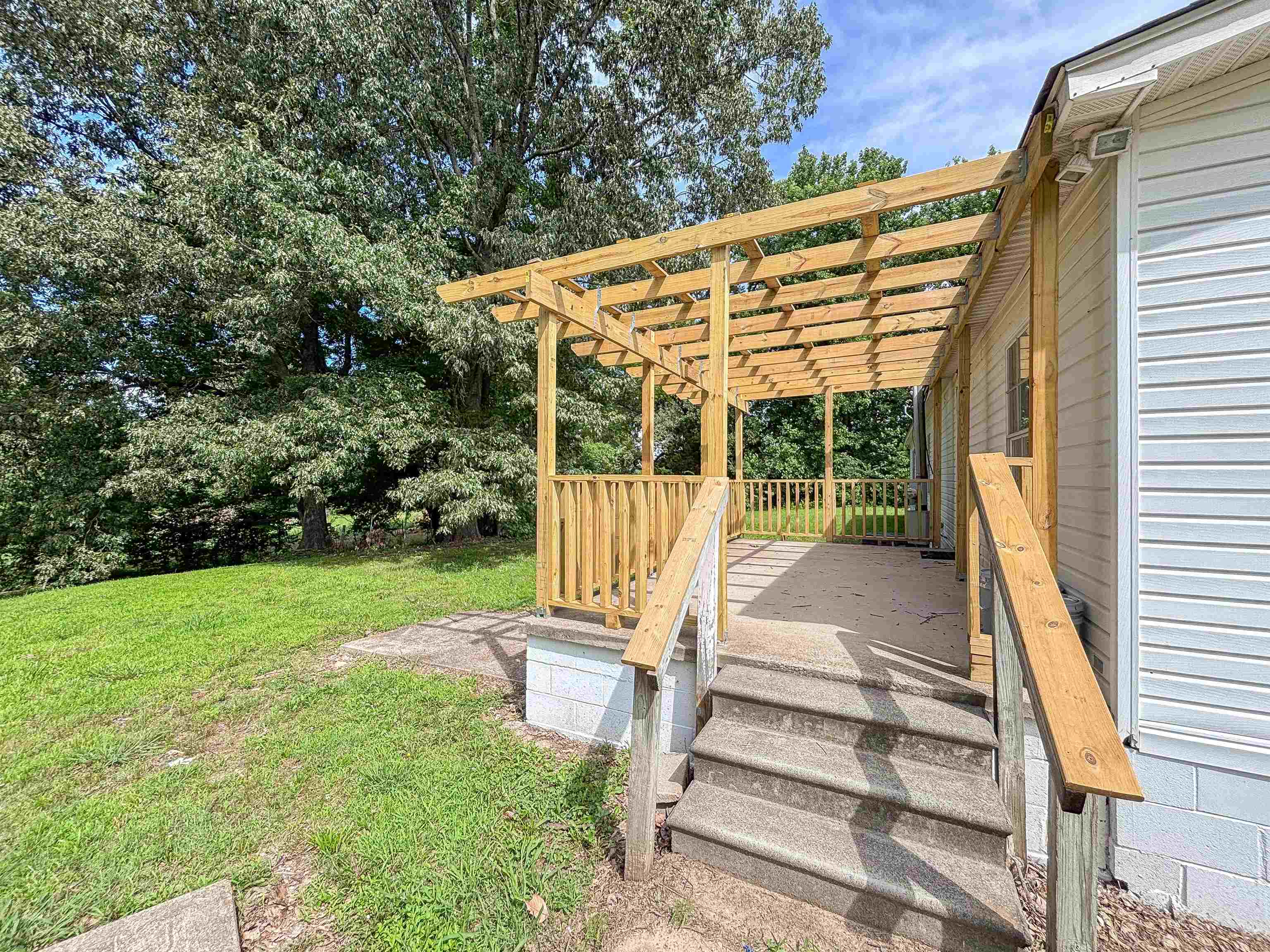 2847 Holly Grove Road Covington, TN 38019 - Photo 33 of 36 Concrete steps leading to an expansive pergola-covered patio with natural wood railings, set against a backdrop of mature trees and a sloped grassy area