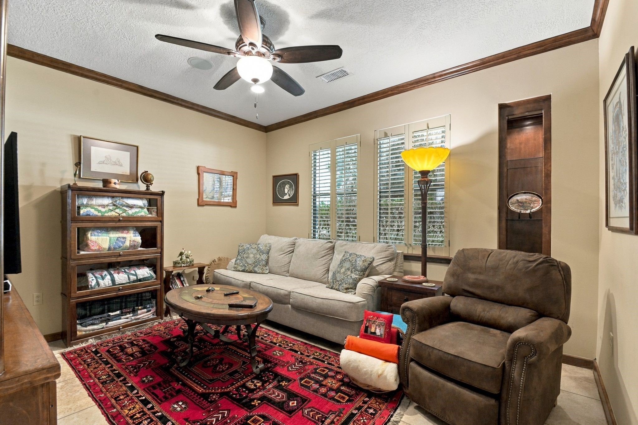 20341 Bay Hill Road Point Blank, TX 77364 - Photo 28 of 50 Cozy secondary bedroom with warm wood accents, featuring a comfortable sofa, plush armchair, and a vibrant red patterned rug. The space is well-lit with natural light from large windows adorned with shutters.
