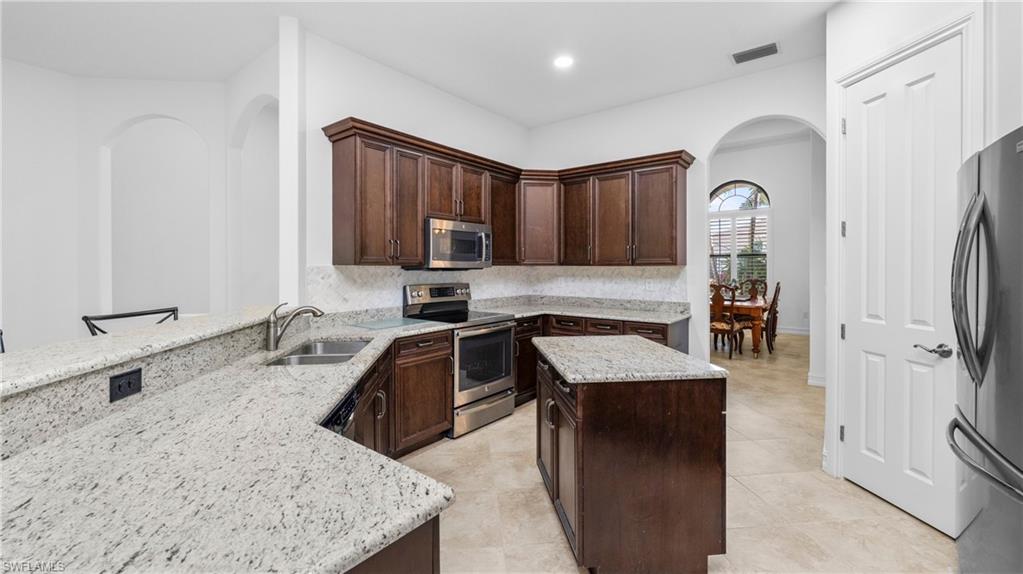8856 Lely Island Circle Naples, FL 34113 - Photo 16 of 50 Kitchen featuring arched walkways, dark wood finish cabinetry, stainless steel appliances, a kitchen island, and light stone counters