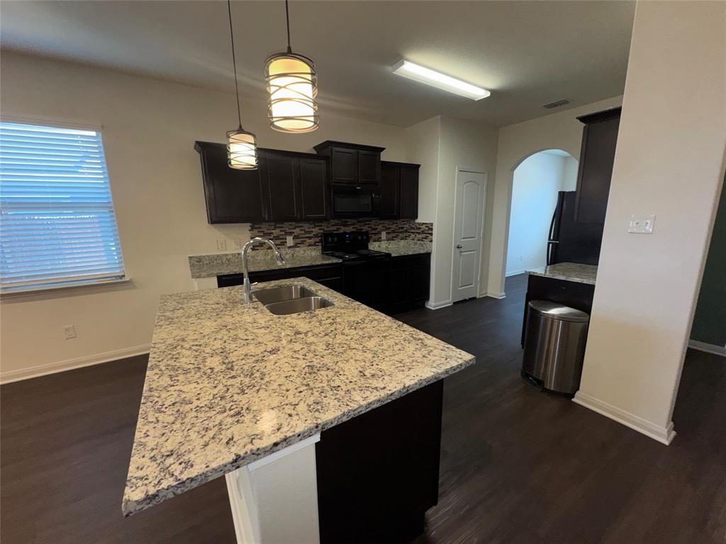 14360 Riata Lane Dallas, TX 75253 - Photo 5 of 16 Kitchen featuring an island with a double basin sink, dark wood-finish cabinetry, light-toned countertops, tile backsplash, and dark wood-finish flooring