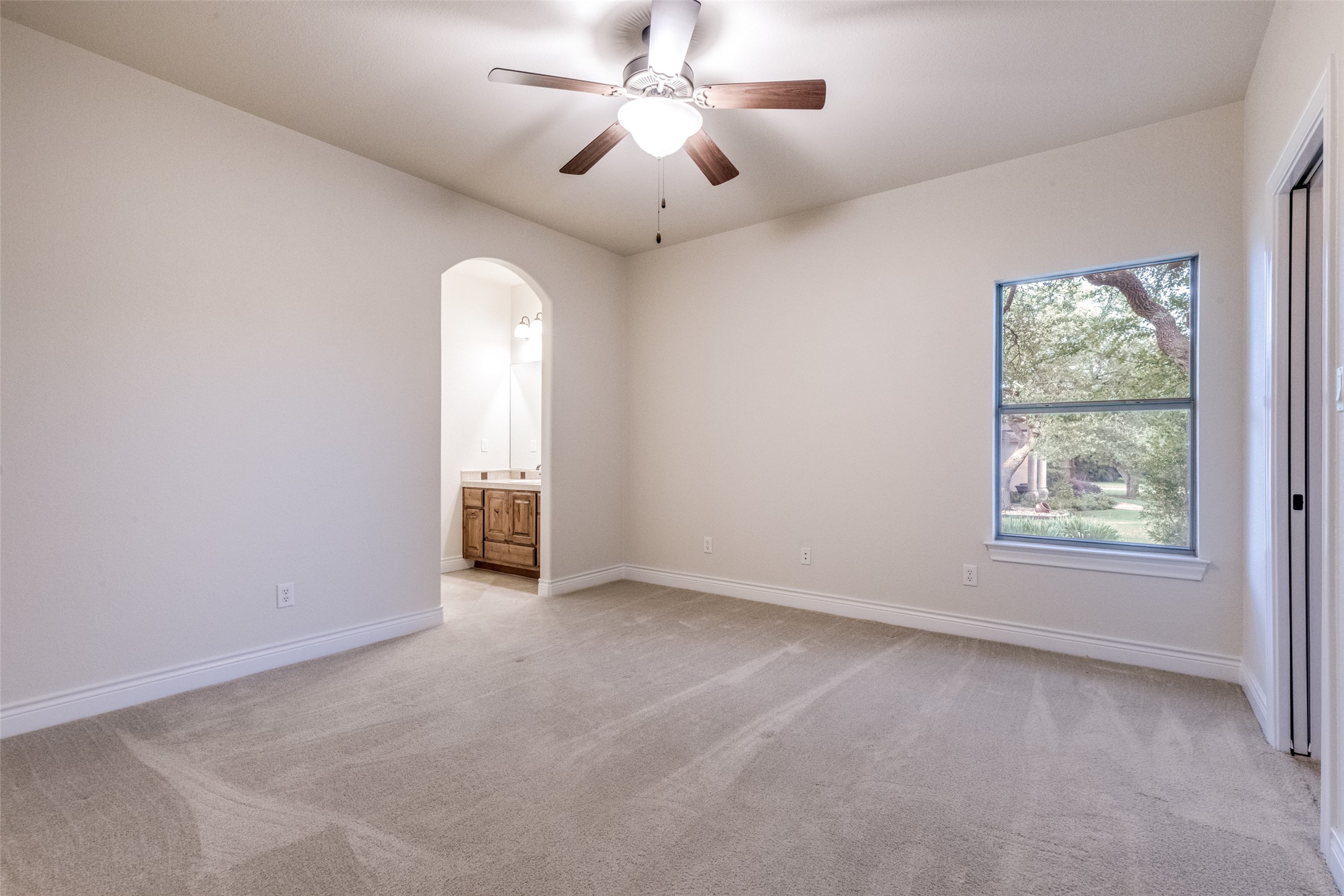 101 Copper Leaf Court Georgetown, TX 78633 - Photo 21 of 31 Spacious carpeted room featuring a ceiling fan with light fixture, a window with exterior views, and an arched entryway to a vanity area with wood cabinetry and a framed mirror