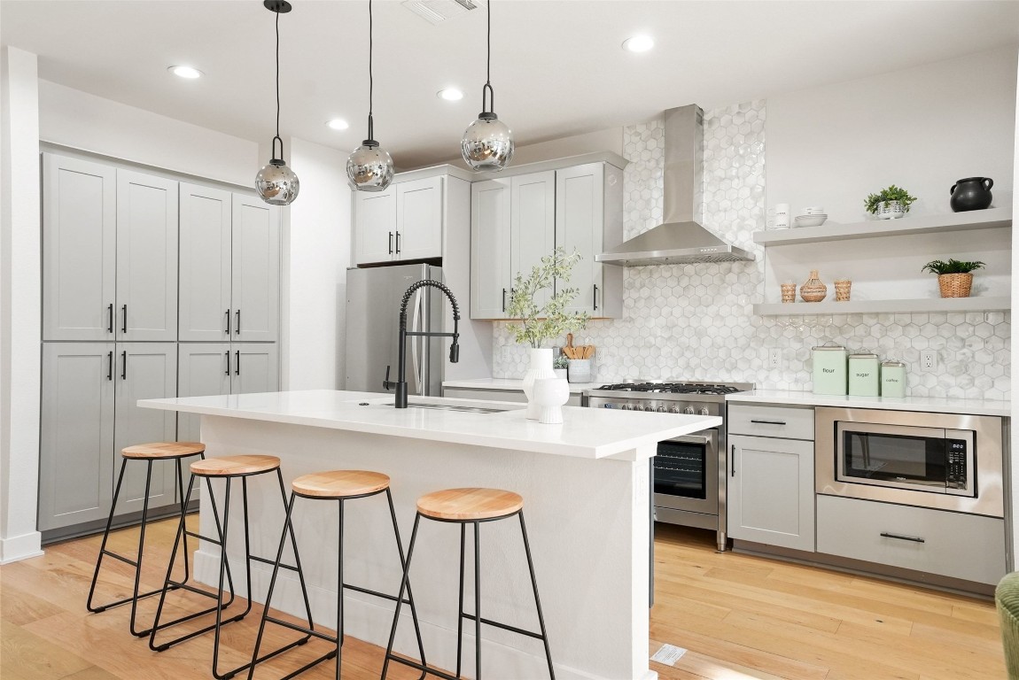 Kitchen with appliances with stainless steel finishes, wall chimney range hood, light wood-style floors, open shelves, and a kitchen breakfast bar
