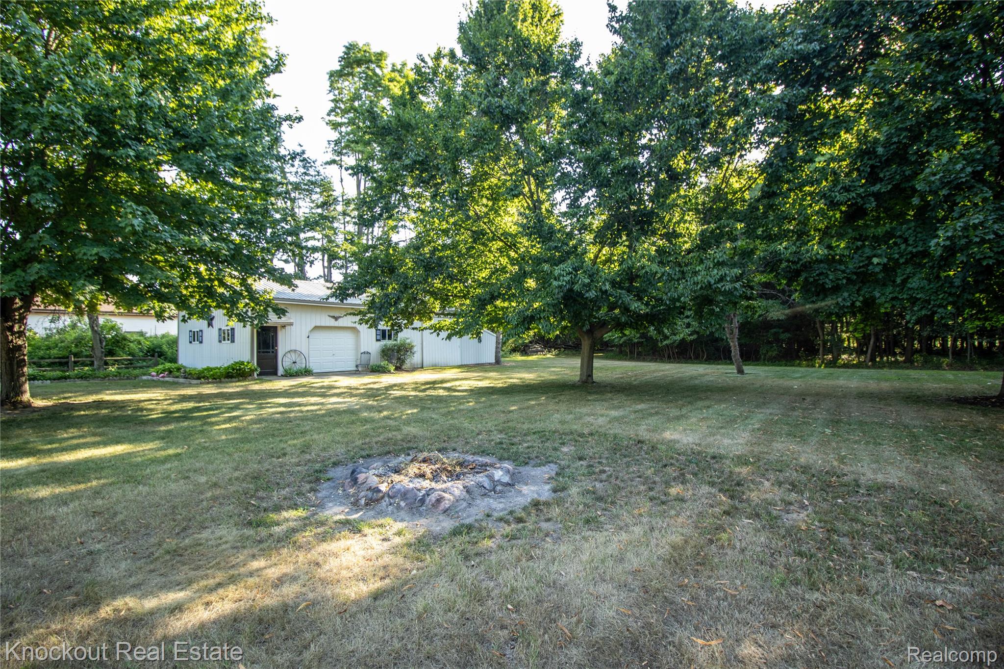 14720 Lincoln Road Chesaning, MI 48616 - Photo 47 of 60 Expansive grassy yard with mature trees, a stone fire pit, and a detached white building featuring a garage door and exterior lighting