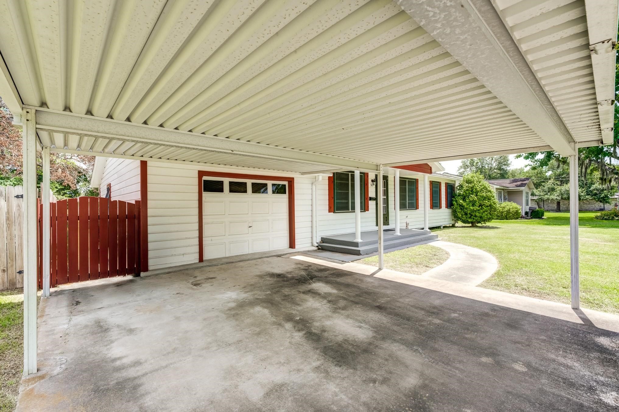 305 Yaupon Street Sweeny, TX 77480 - Photo 7 of 22 This photo shows a covered carport leading to a single-car garage attached to a charming house with a well-maintained lawn and a wooden fence. The exterior features white siding with red accents, creating a welcoming appearance.