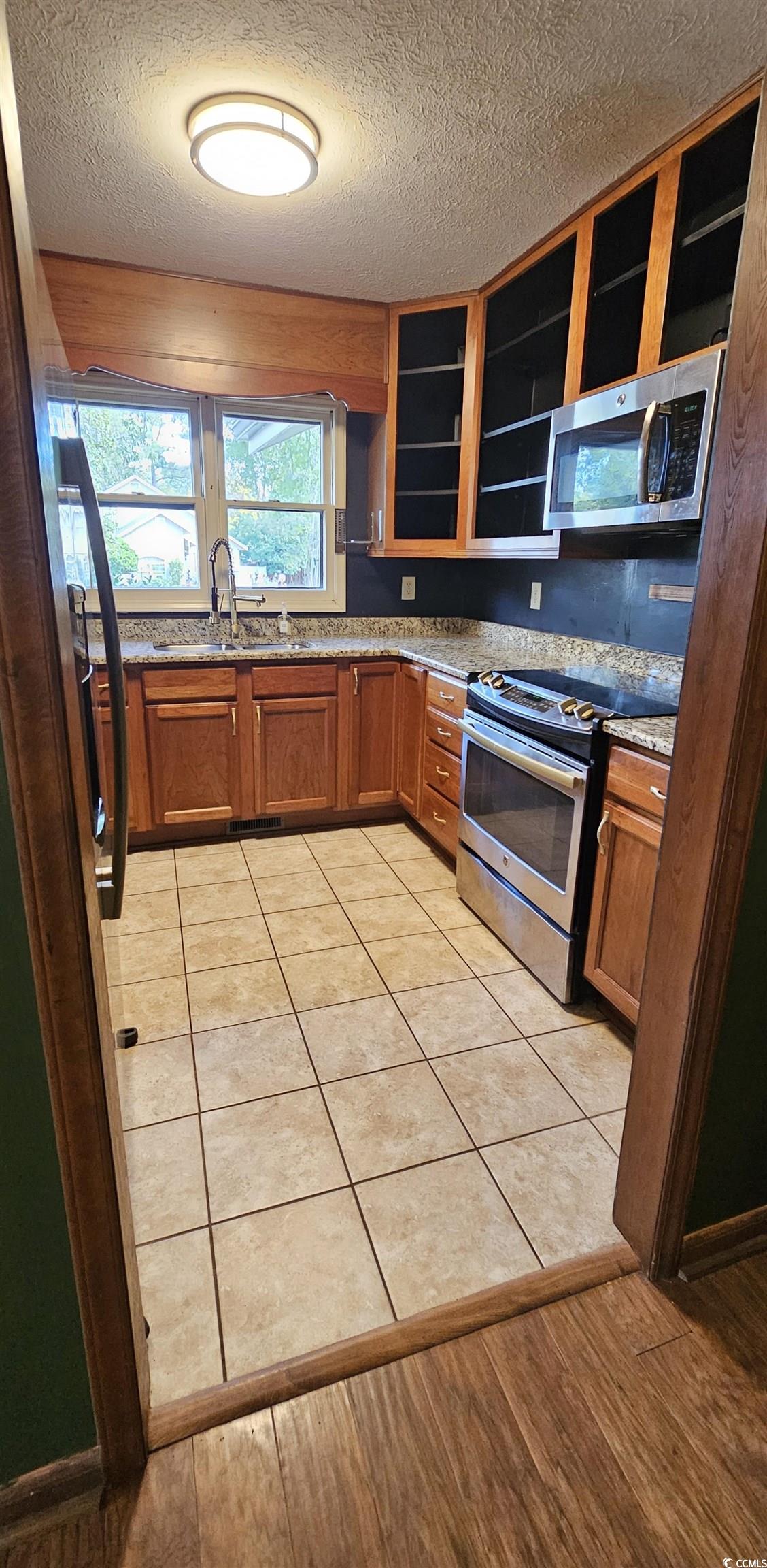 117 Partridge Berry Road Myrtle Beach, SC 29579 - Photo 6 of 33 Kitchen with brown cabinetry, appliances with stainless steel finishes, a textured ceiling, light stone counters, and light tile patterned floors