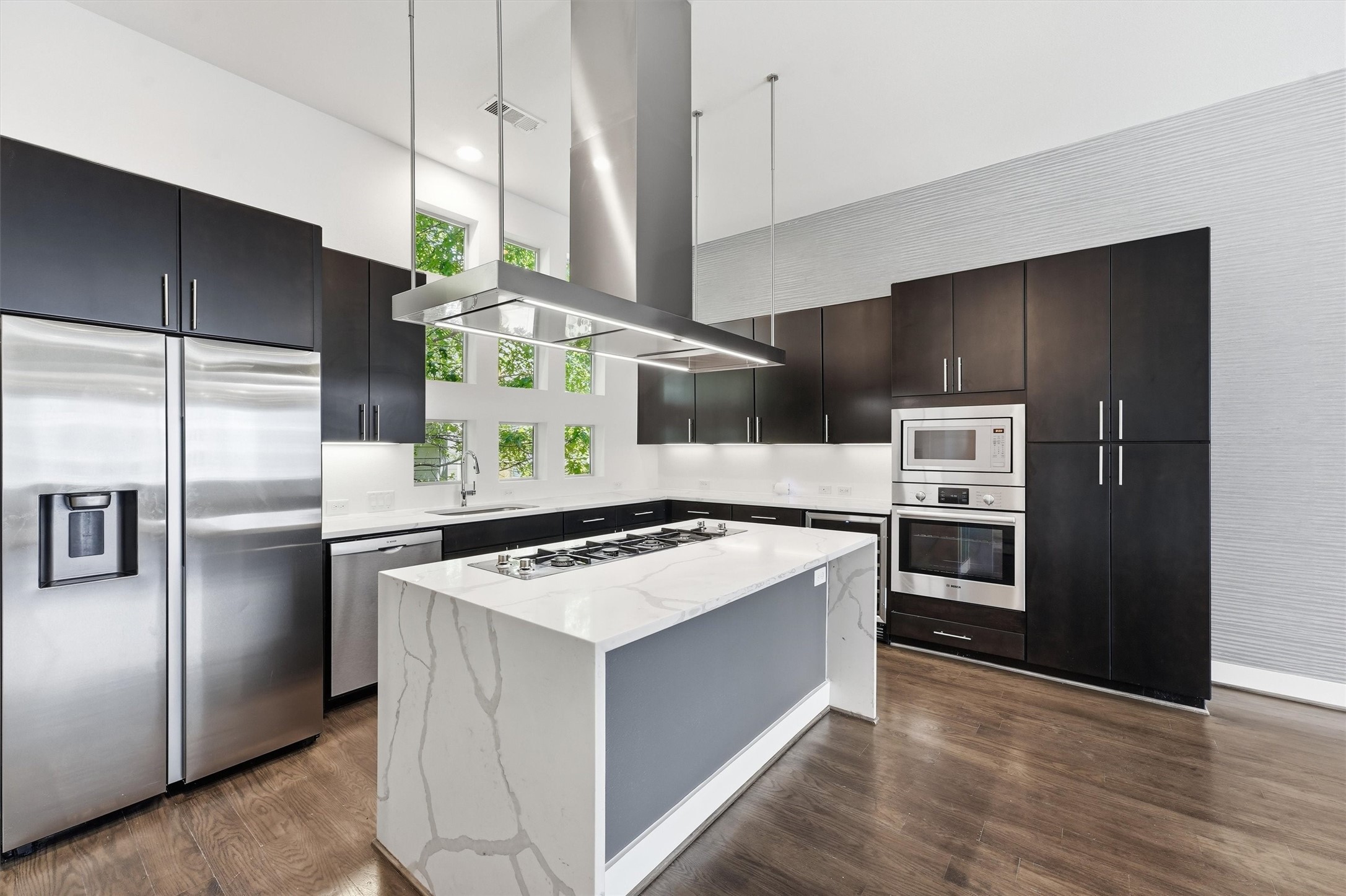 2338 Commerce Street Houston, TX 77003 - Photo 1 of 29 This modern kitchen features sleek dark cabinets, stainless steel appliances, and a large quartz waterfall island with a gas cooktop. The space is brightened by high ceilings and a row of windows, offering a contemporary and functional design.