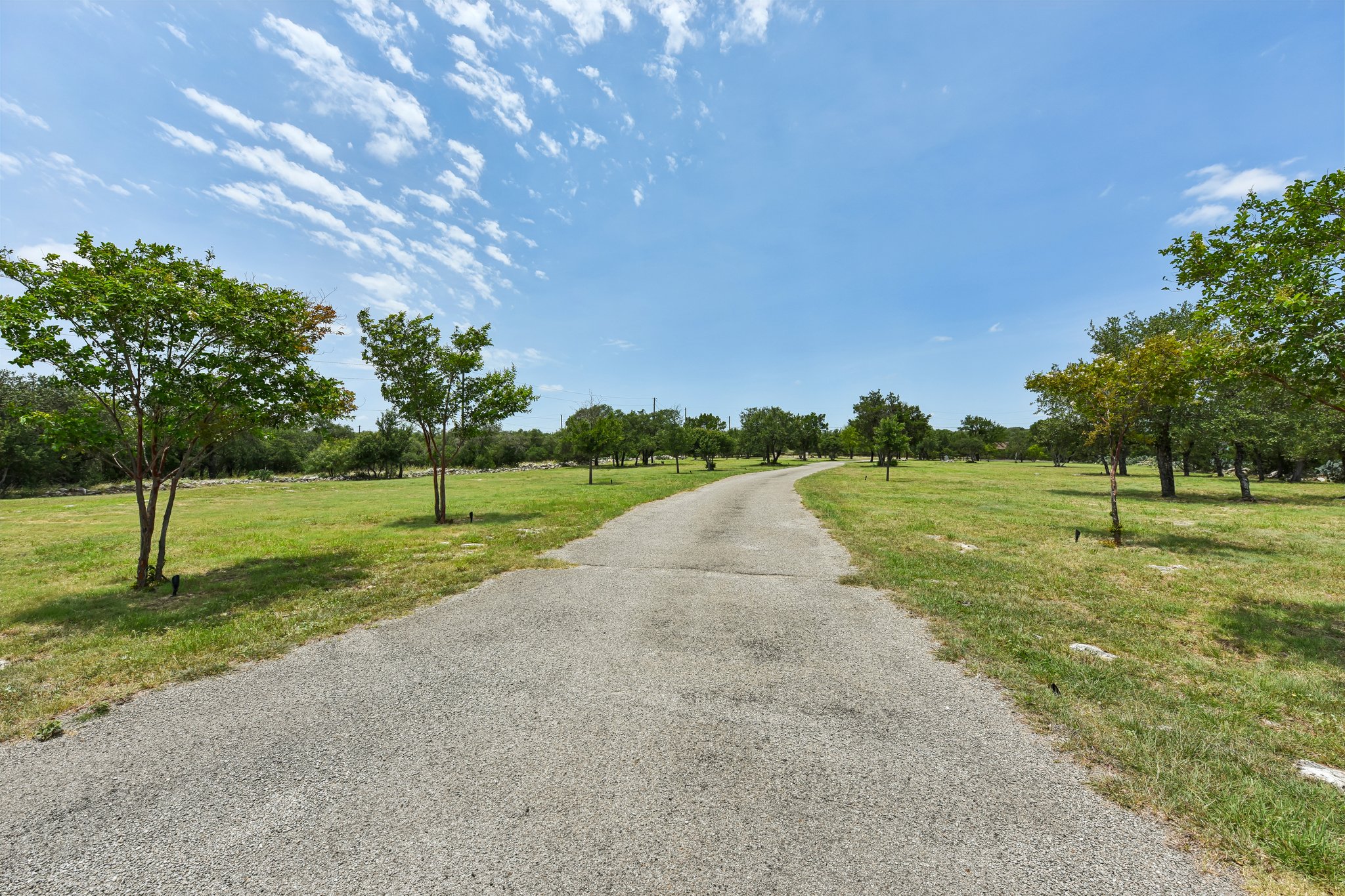 2203 Improver Road Spicewood, TX 78669 - Photo 25 of 38 A long paved driveway winds through lush green grounds with mature trees under a clear blue sky.