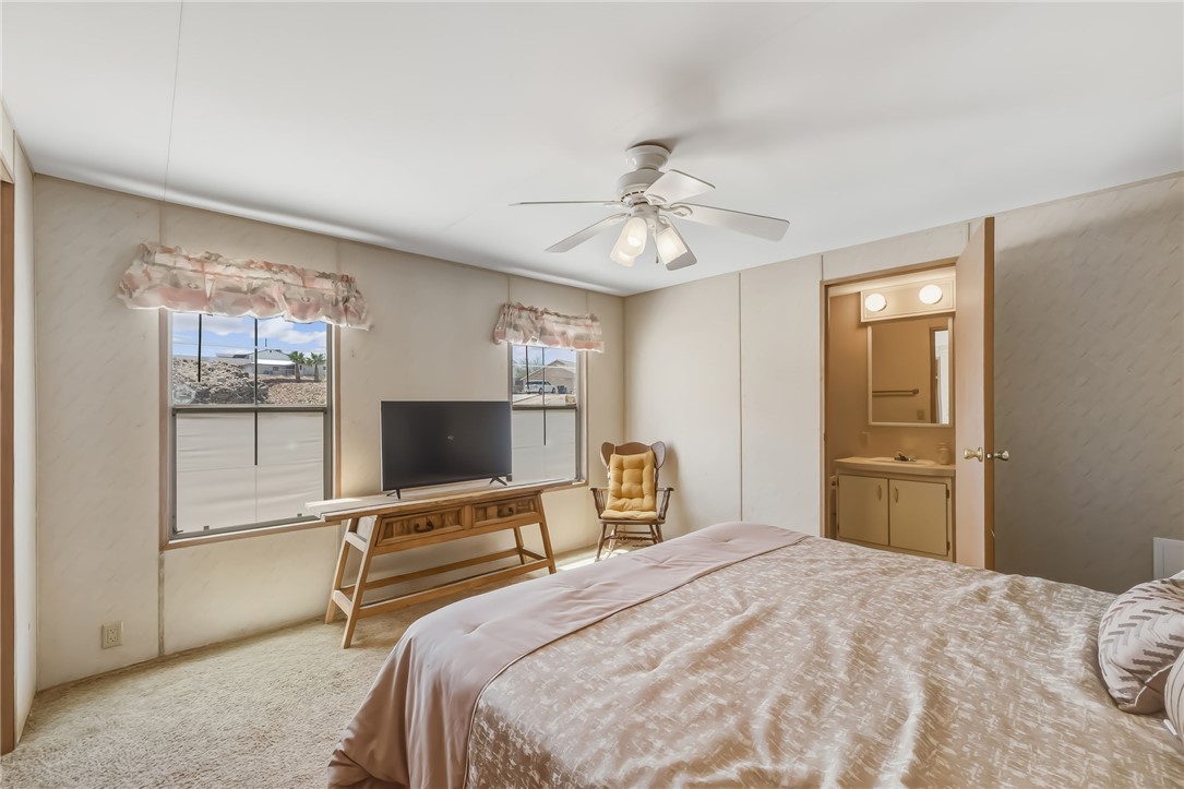 3880 Rising Sun Road Bullhead City, AZ 86442 - Photo 25 of 42 Carpeted bedroom with two windows, a ceiling fan with light fixture, and an open doorway revealing a vanity with a sink and mirror