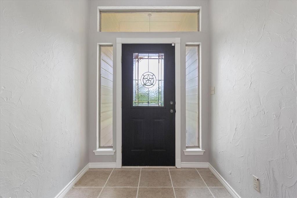 140 Oak Ridge Loop Whitney, TX 76692 - Photo 4 of 40 The property features a front entry with a dark-colored door, flanked by narrow vertical windows and topped with a horizontal transom window, all set against light-colored walls and tiled flooring