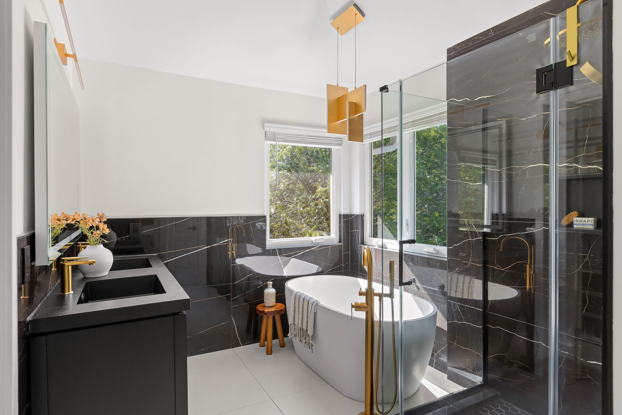 6500 Clay Allison Pass Austin, TX 78749 - Photo 24 of 39 Modern bathroom featuring a freestanding soaking tub with gold-finish floor-mount filler, an enclosed shower with glass doors and dark marble-look tile, a dual vanity with integrated black sinks and gold-finish faucets, and an abstract gold-finish pendant light fixture