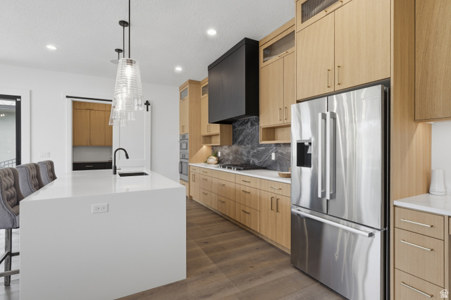 653 East Vandalay Lane Draper, UT 84020 - Photo 20 of 61 Modern kitchen featuring a white center island with an undermount sink and matte black faucet, wood-finish cabinetry, stainless steel appliances, a black range hood, and wood-finish flooring