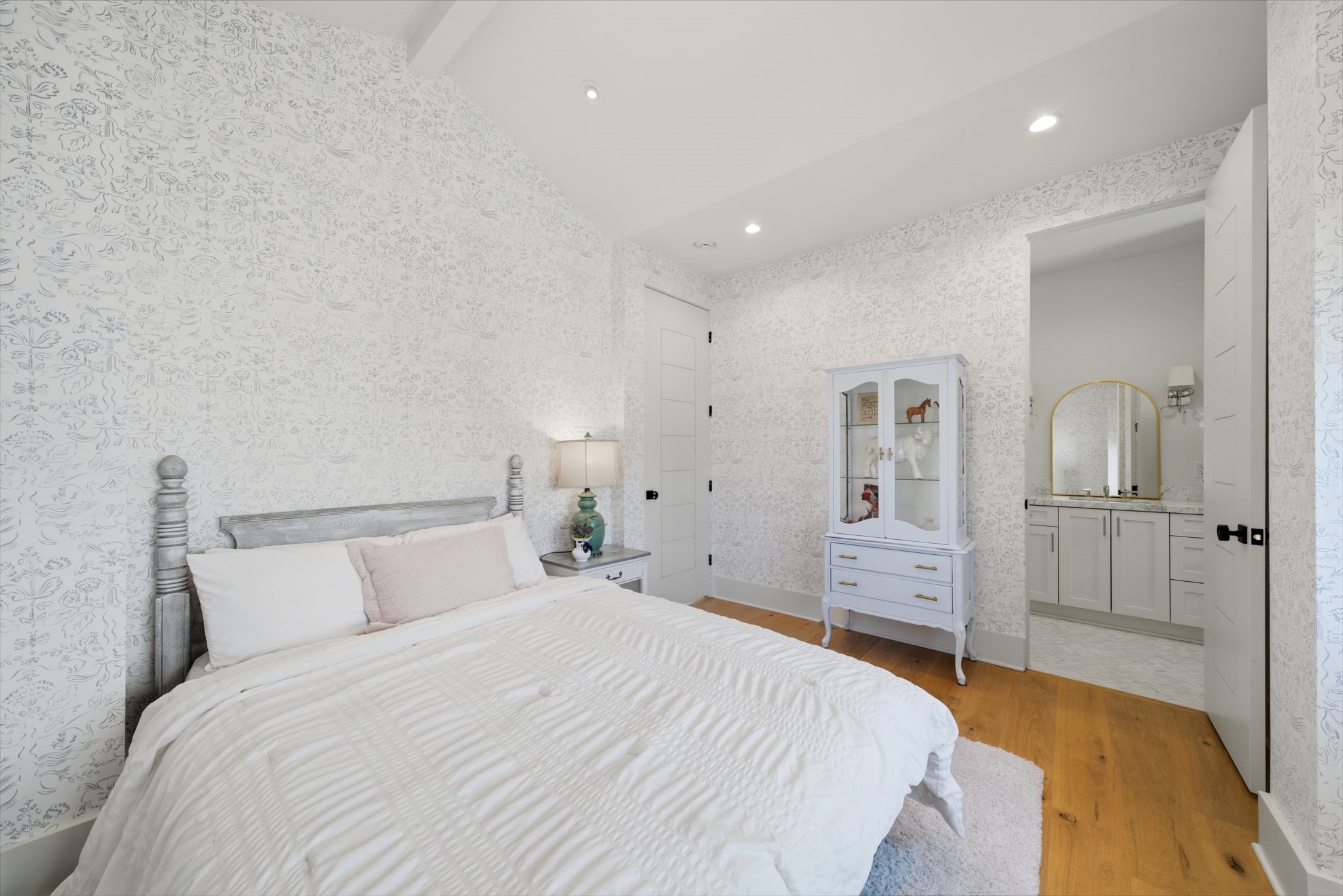 2615 Yupon Street Houston, TX 77006 - Photo 37 of 50 Delicate floral designer wallpaper and vaulted ceilings create a sophisticated atmosphere in this light-filled secondary bedroom. Wide-plank white oak floors lead to a private ensuite bath, while a white glass-front armoire adds elegant storage. Recessed LED lighting and a neutral palette enhance the contemporary comfort of this refined retreat.