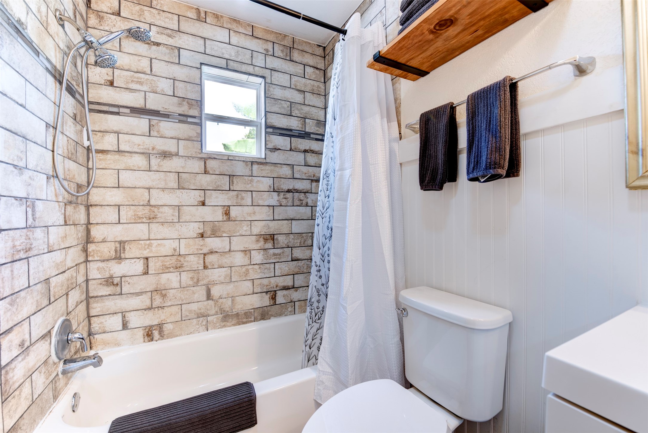 1121 Panama Street Houston, TX 77009 - Photo 22 of 35 Tub and shower detail featuring floor-to-ceiling rustic brick-style large-format tile, chrome handheld shower head with slide bar, white soaking tub, operable window for natural light and ventilation, natural wood floating shelf with black iron rod, and beadboard wainscoting.