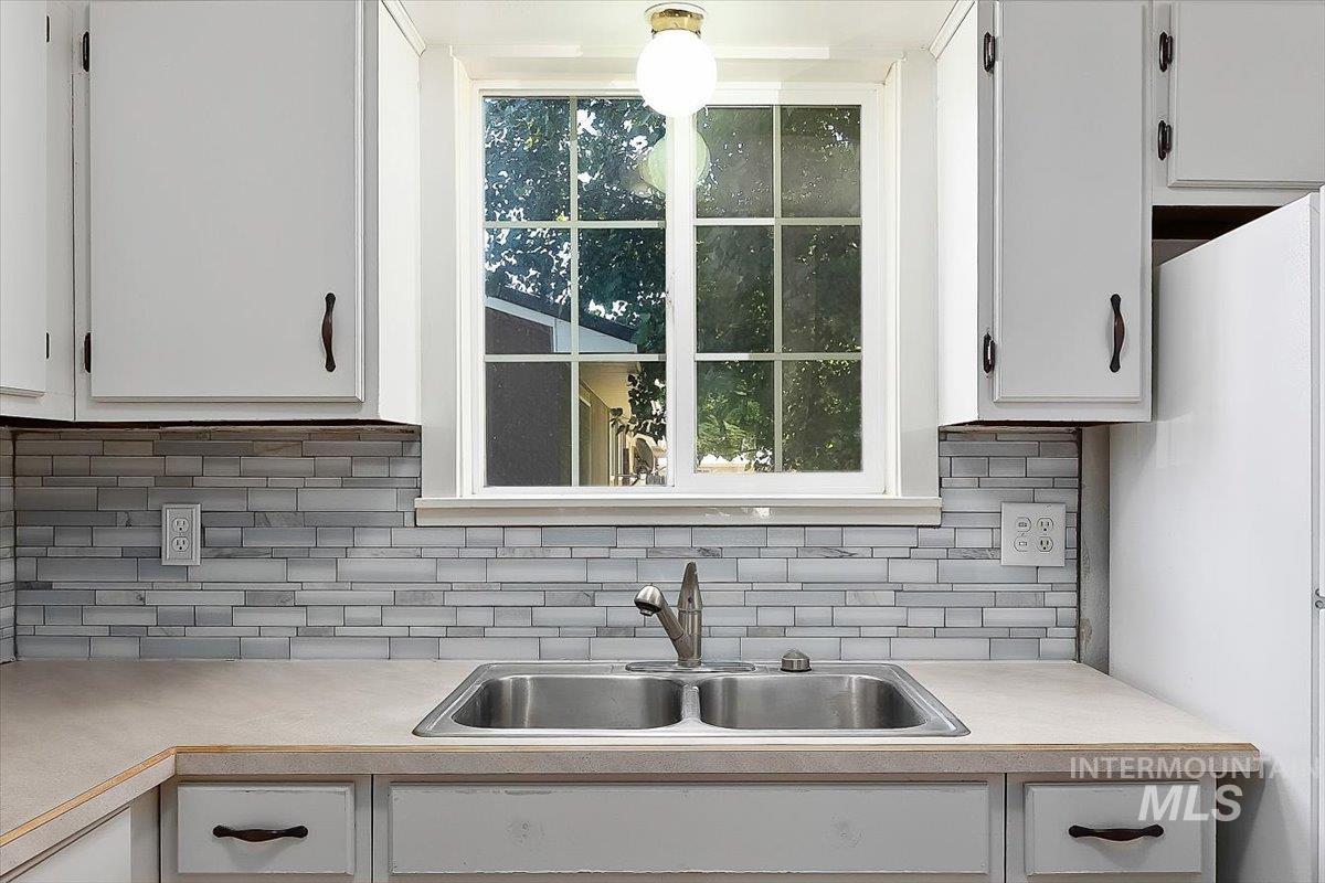 1940 Q Street Heyburn, ID 83336 - Photo 17 of 30 Kitchen featuring a double basin stainless steel sink, a window with grid panes, white cabinetry with dark hardware, a mosaic tile backsplash, and light-toned countertops