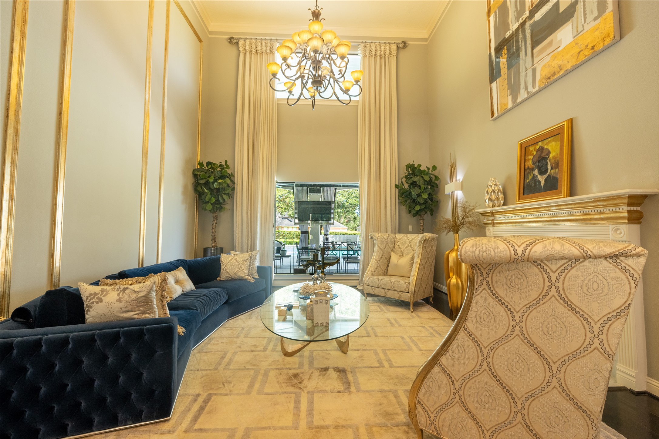 4002 Silver Ridge Boulevard Missouri City, TX 77459 - Photo 11 of 50 This elegant living space features high ceilings with classic detailing, a sophisticated chandelier, and full-length windows leading to a balcony. The room is furnished with a plush blue sofa, ornate armchairs, and a glass coffee table, all complemented by tasteful artwork and decor.