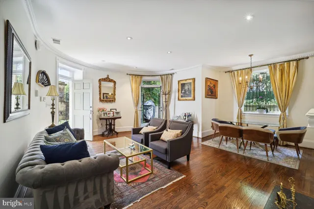a living room with furniture two large window and wooden floor