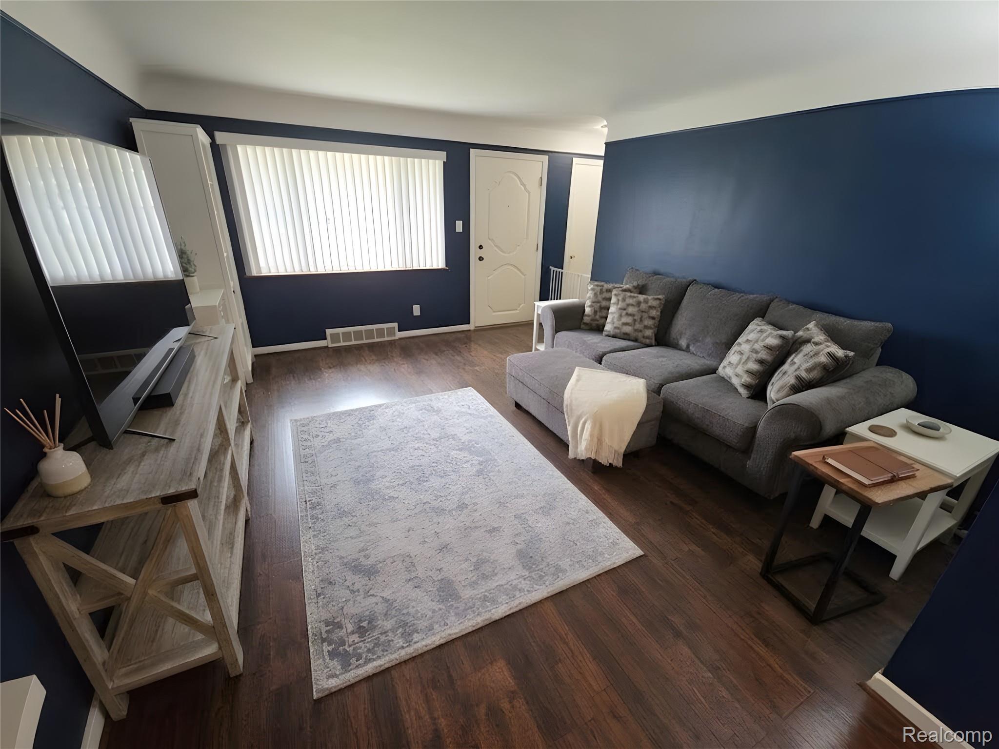 9921 Middlebelt Road, Unit 32 Livonia, MI 48150 - Photo 3 of 11 Living area featuring dark wood-finish flooring, a white entry door, and a prominent window with vertical blinds