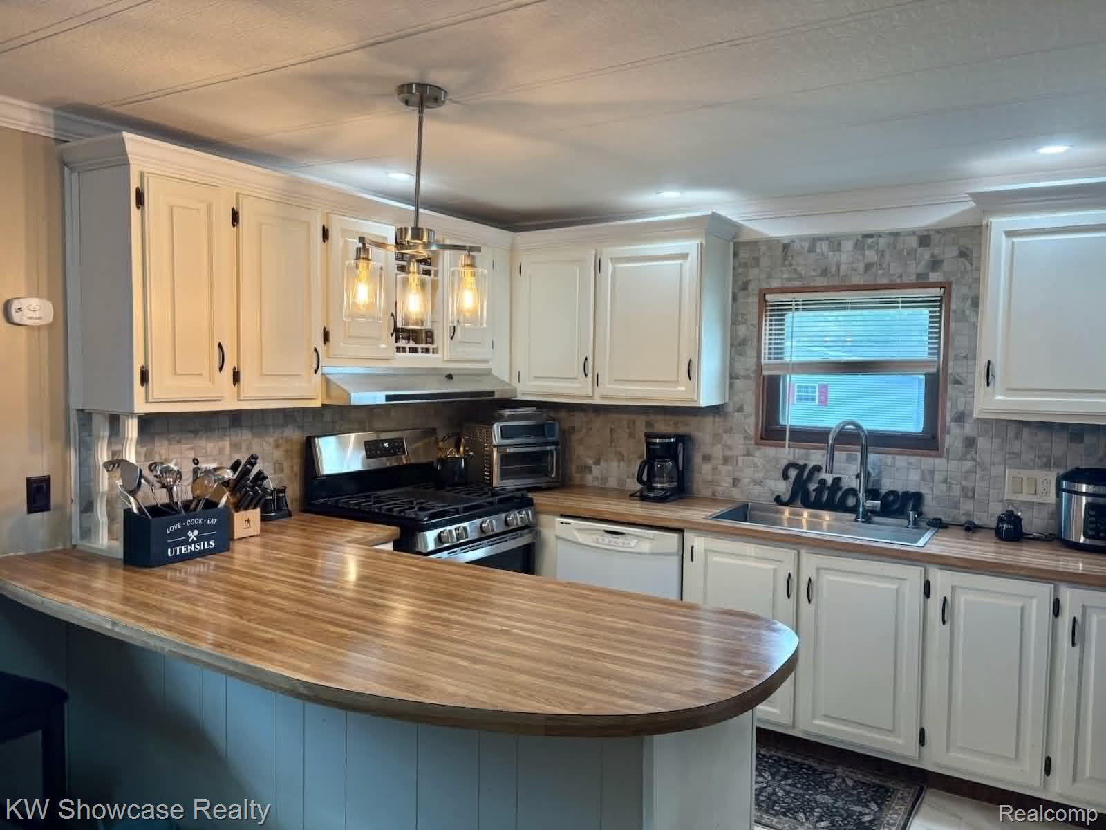 9435 Hunter Rills Road Fenton, MI 48430 - Photo 2 of 13 Thoughtfully renovated kitchen featuring cream-painted cabinetry, wood-finish countertops with rounded island seating, stainless steel appliances, tile backsplash, pendant lighting, and abundant natural light from multiple windows