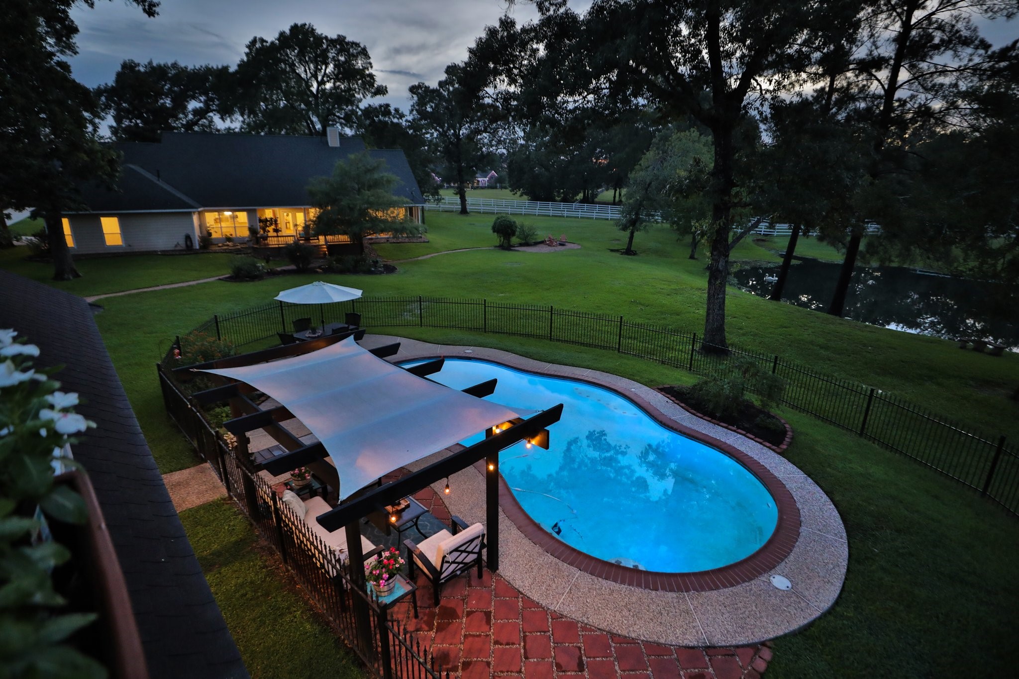 34310 High Point Drive Magnolia, TX 77355 - Photo 34 of 48 This aerial view shows a spacious backyard with a serene pool, shaded pergola area, and lush greenery, ideal for relaxation and entertaining. The house is warmly lit and offers a picturesque view of the pond and surrounding trees, creating a tranquil and inviting atmosphere.