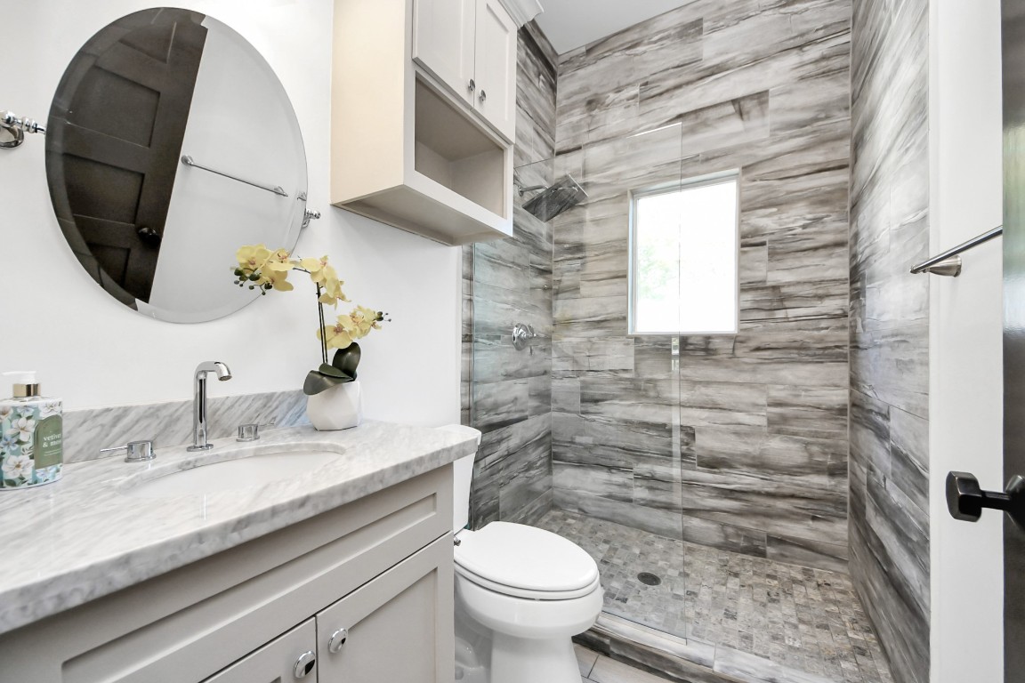 1202 Archer Street Houston, TX 77009 - Photo 36 of 47 This modern bathroom located on the ground level offers a sleek design with a round mirror, a marble countertop, and a frameless-enclosed shower. The shower area has stylish wood-look tiles and a small window providing natural light.