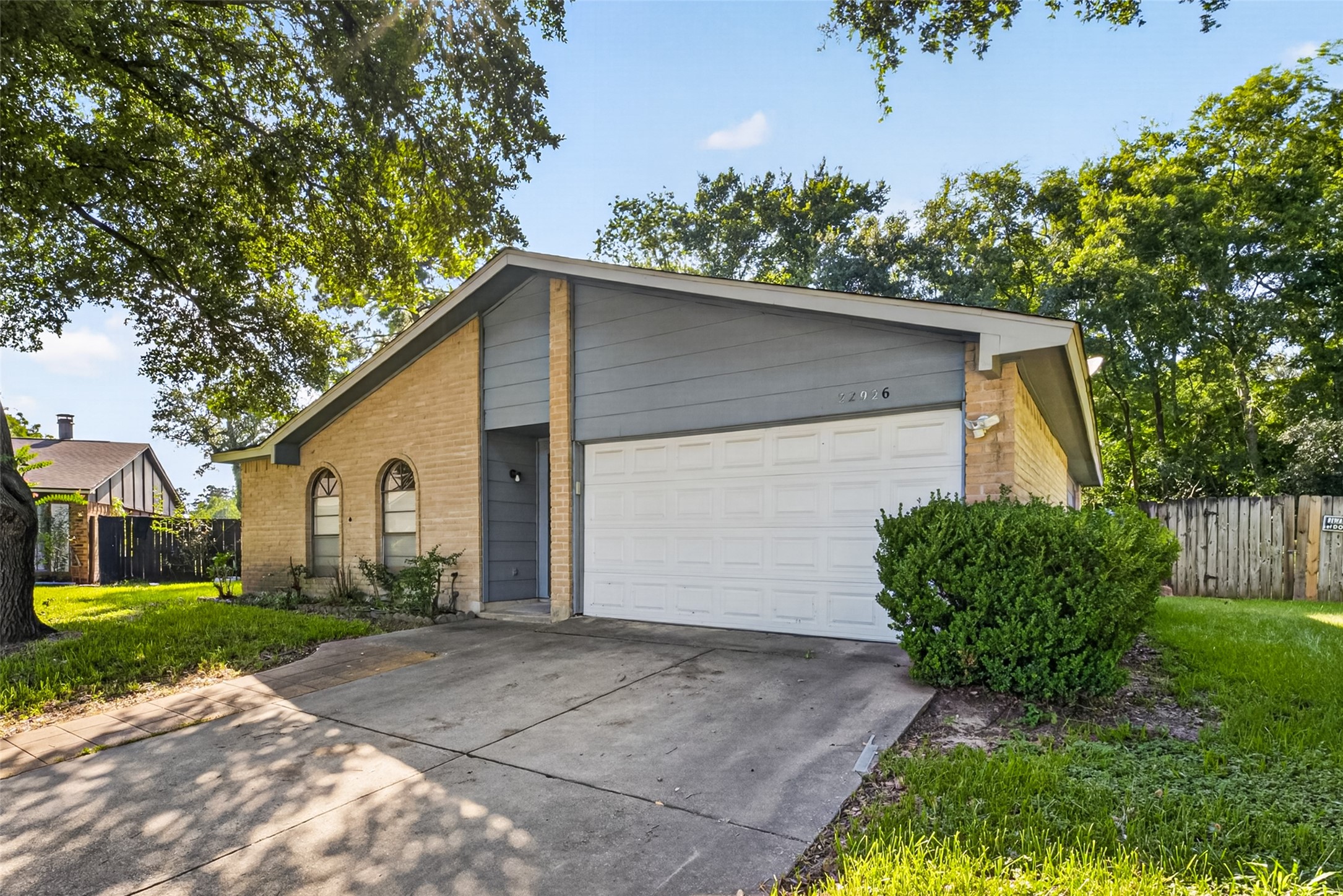 A charming single-story home with a two-car garage and a sloped roof, set on a lush green lot. The exterior features a combination of brick and siding, with mature trees providing shade. Perfect for comfortable living in a serene neighborhood.
