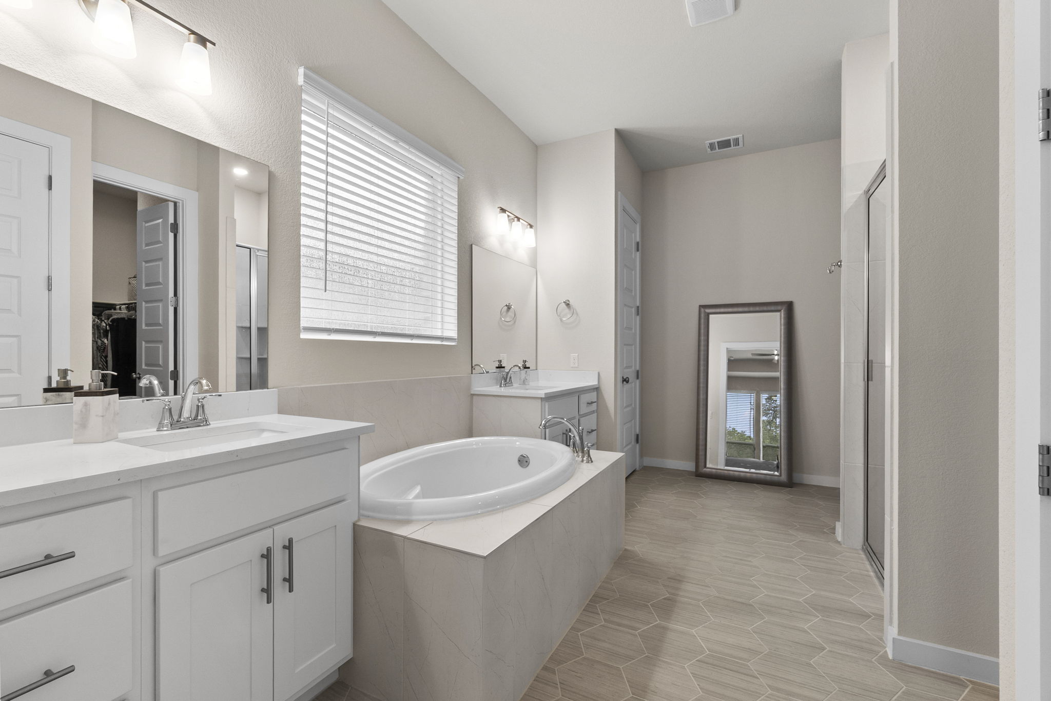 460 Constitution Way Kyle, TX 78640 - Photo 20 of 40 Expansive bathroom featuring dual white vanities with integrated sinks, a built-in soaking tub with surrounding tile, a walk-in shower with a glass enclosure, and hexagonal wood-finish flooring