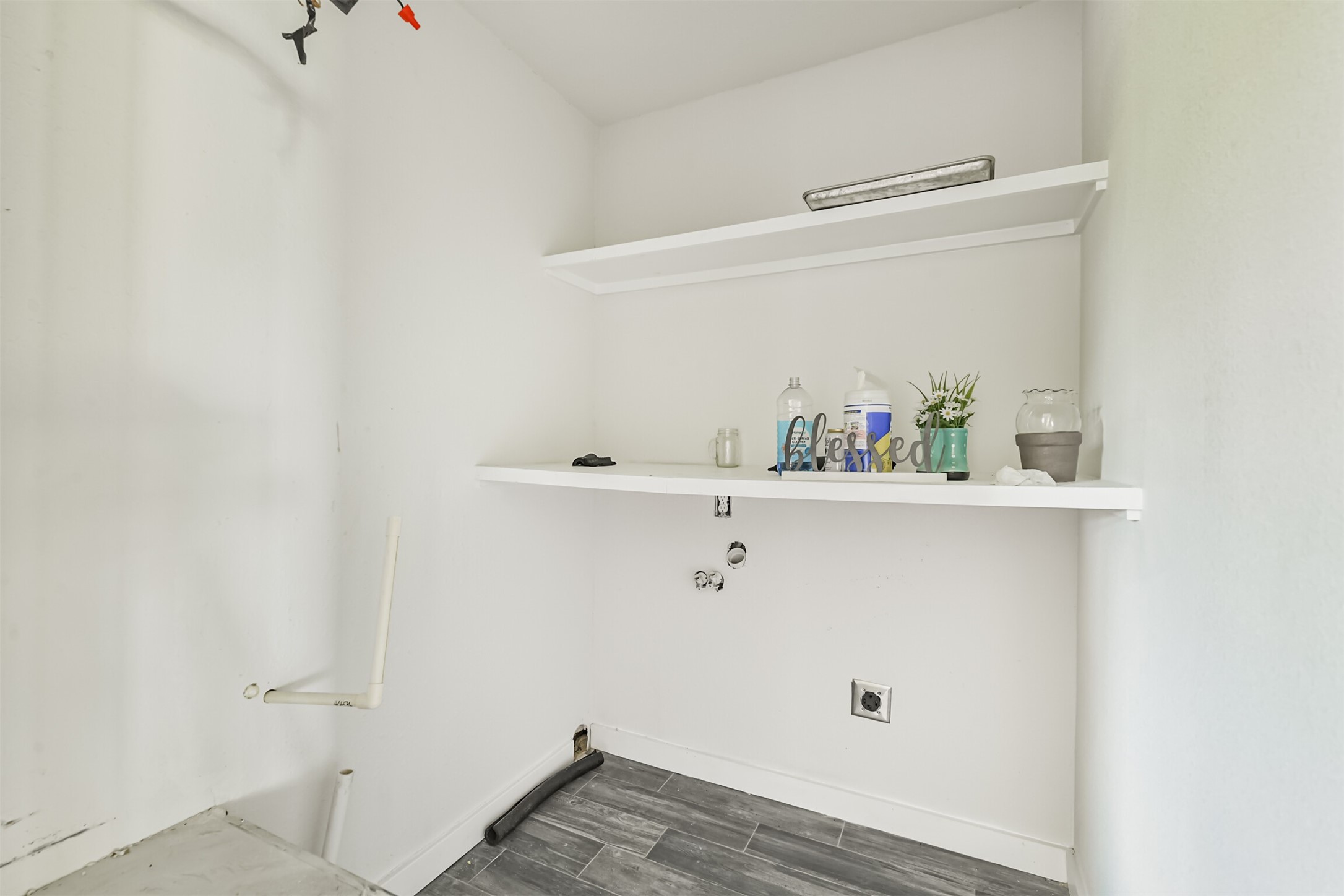 209 Leago Street Houston, TX 77022 - Photo 10 of 24 This photo shows a utility room with plumbing hookups, ideal for a washer and dryer. It features sleek gray tile flooring, white walls, and two white shelves for storage.