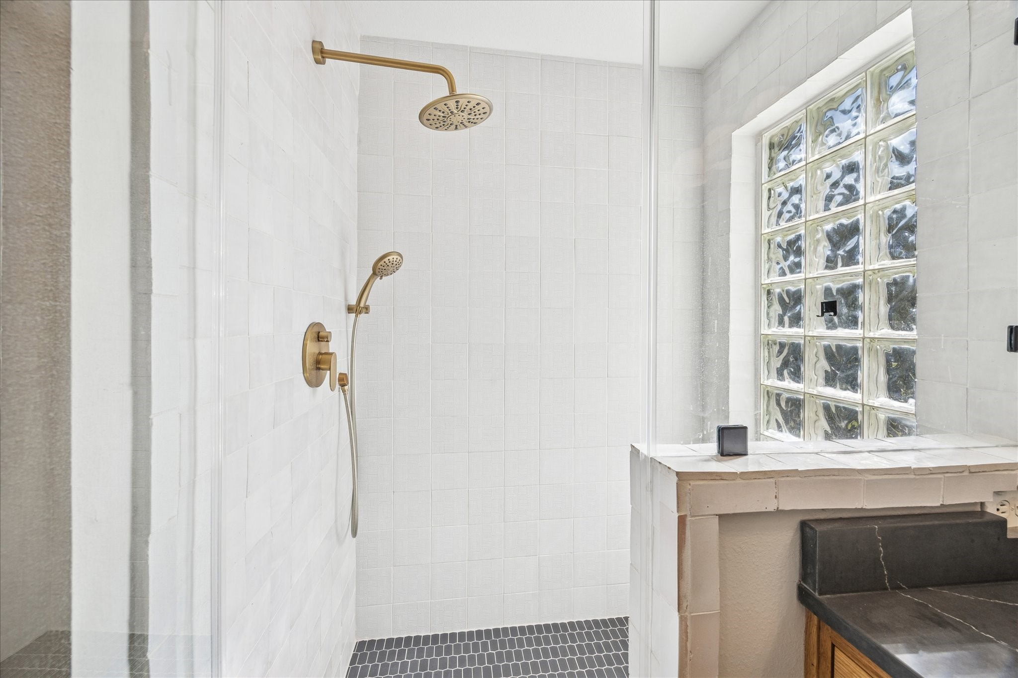 2607 Link Road Houston, TX 77009 - Photo 18 of 21 Primary Bathroom shower with elegant gold fixtures, white tiled walls, and a glass block window for natural light. The space is complemented by a dark countertop, adding a touch of sophistication.
