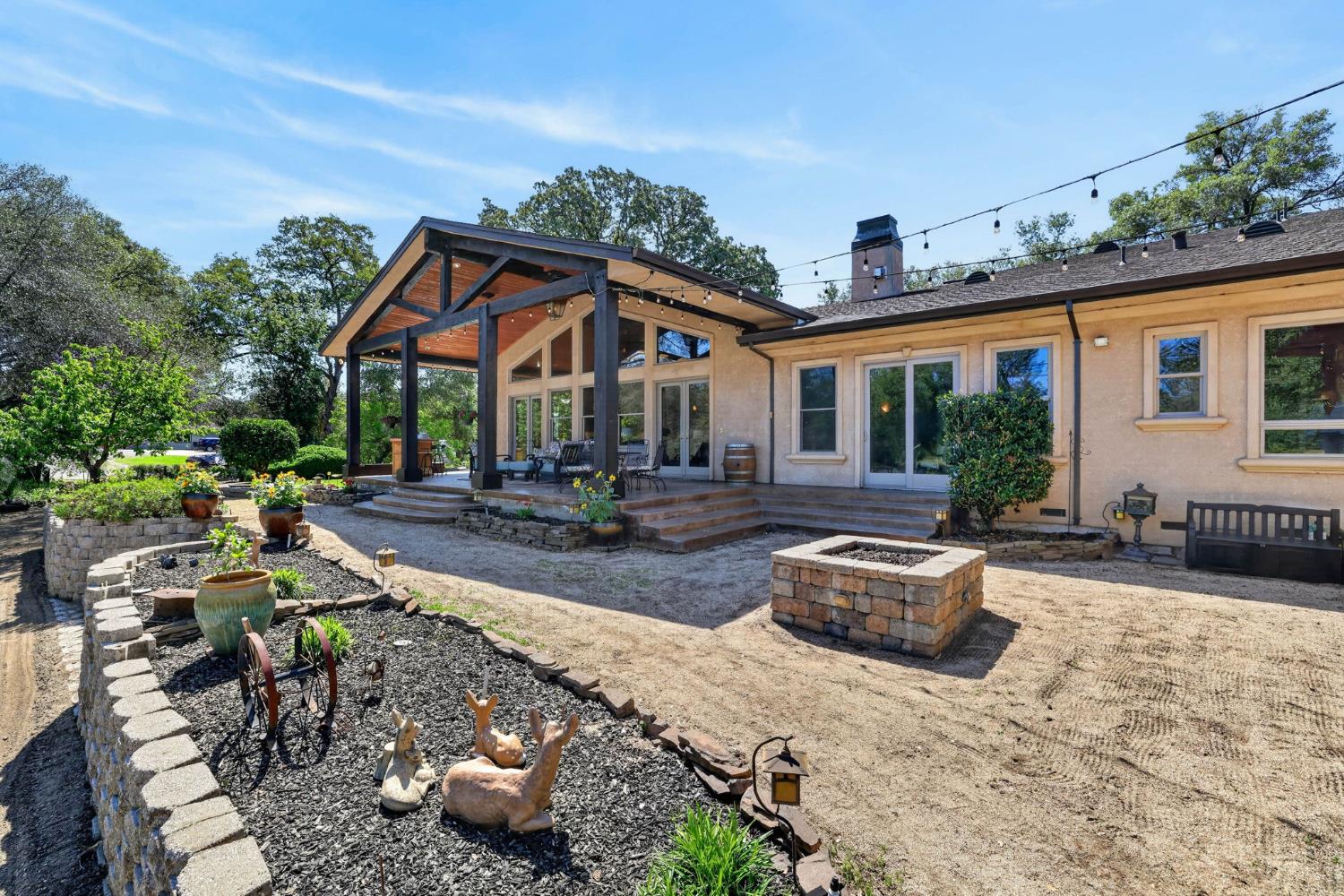 7021 Horseshoe Bar Road Loomis, CA 95650 - Photo 53 of 77 Expansive rear exterior featuring a covered patio with exposed beam ceiling, multiple glass doors, a stone-clad fire pit, tiered landscaping with stone retaining walls, and string lighting