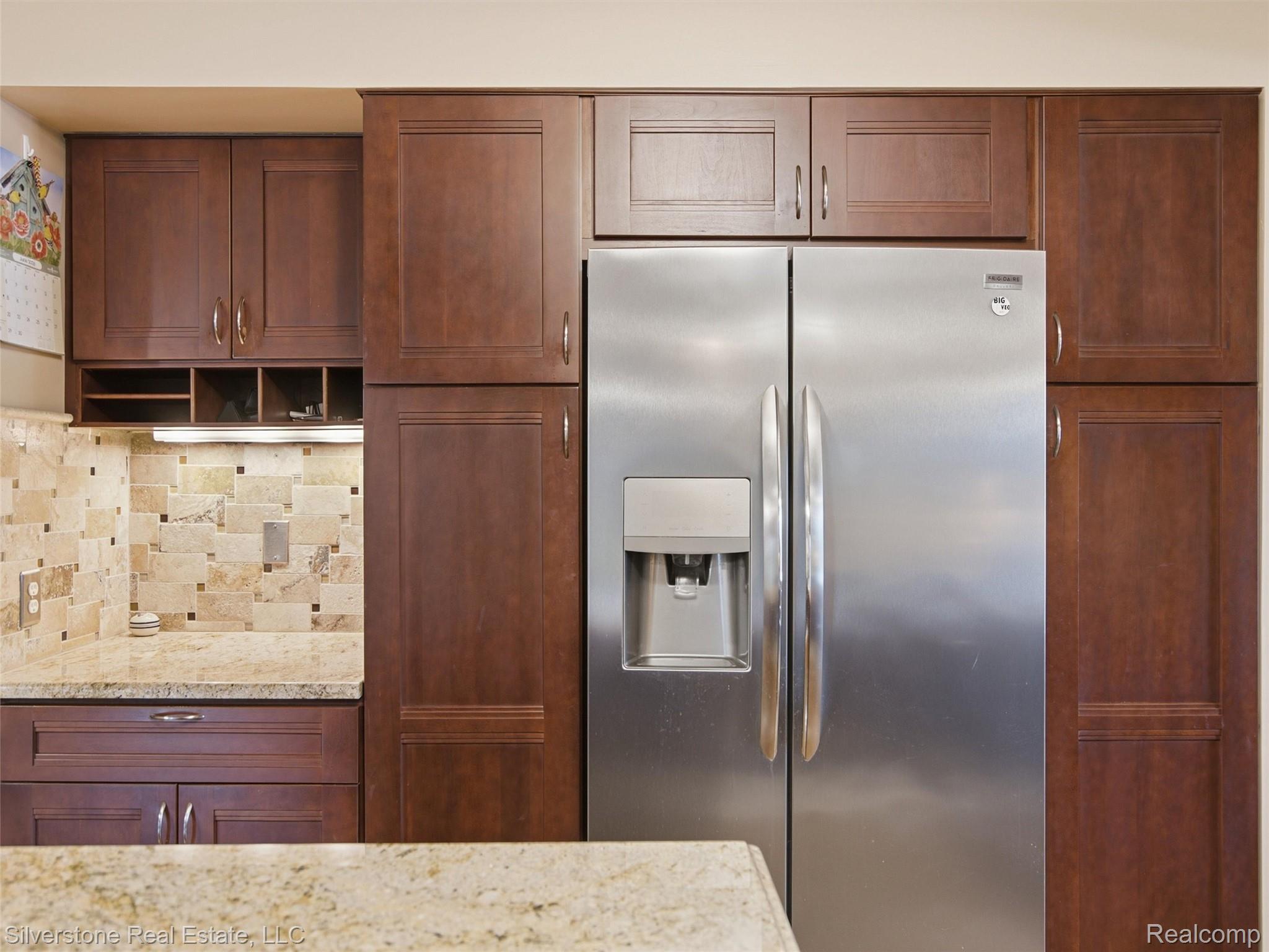 6577 Edith Court Troy, MI 48085 - Photo 26 of 61 Stainless steel side-by-side refrigerator, rich wood-finish cabinetry, under-cabinet lighting, natural stone tile backsplash, and light-toned countertop