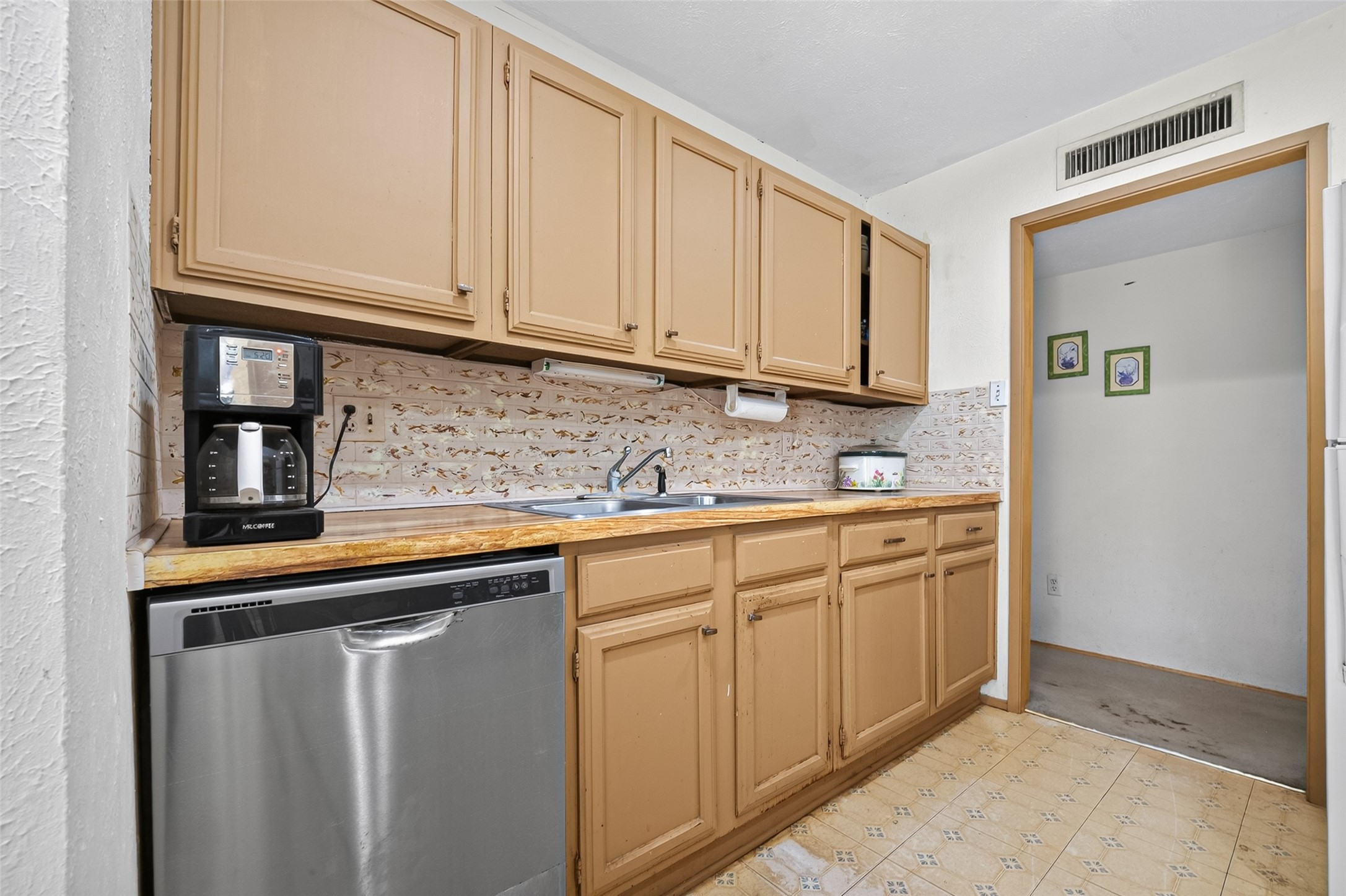 22926 Millgate Drive Spring, TX 77373 - Photo 13 of 34 Cozy kitchen with warm-toned cabinets and patterned backsplash. Features include a stainless steel dishwasher, a coffee maker, and ample countertop space. The room opens to an adjacent area with decorative wall art.
