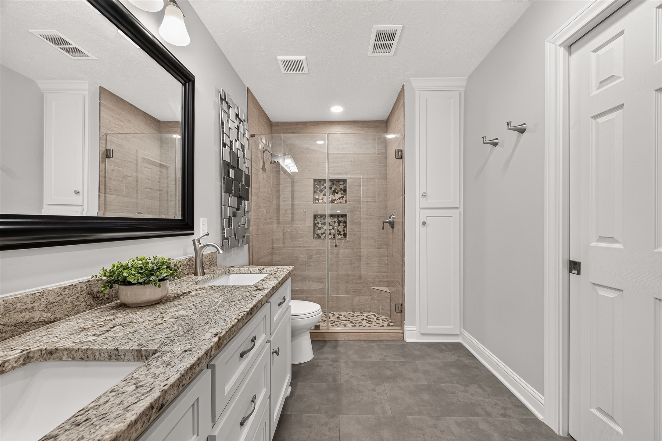 24719 Wilderness Road Spring, TX 77380 - Photo 29 of 42 This primary bathroom features a modern design with a granite countertop, large mirror, and sleek fixtures. It includes a spacious glass-enclosed shower with stylish tile work and ample storage with white cabinetry.
