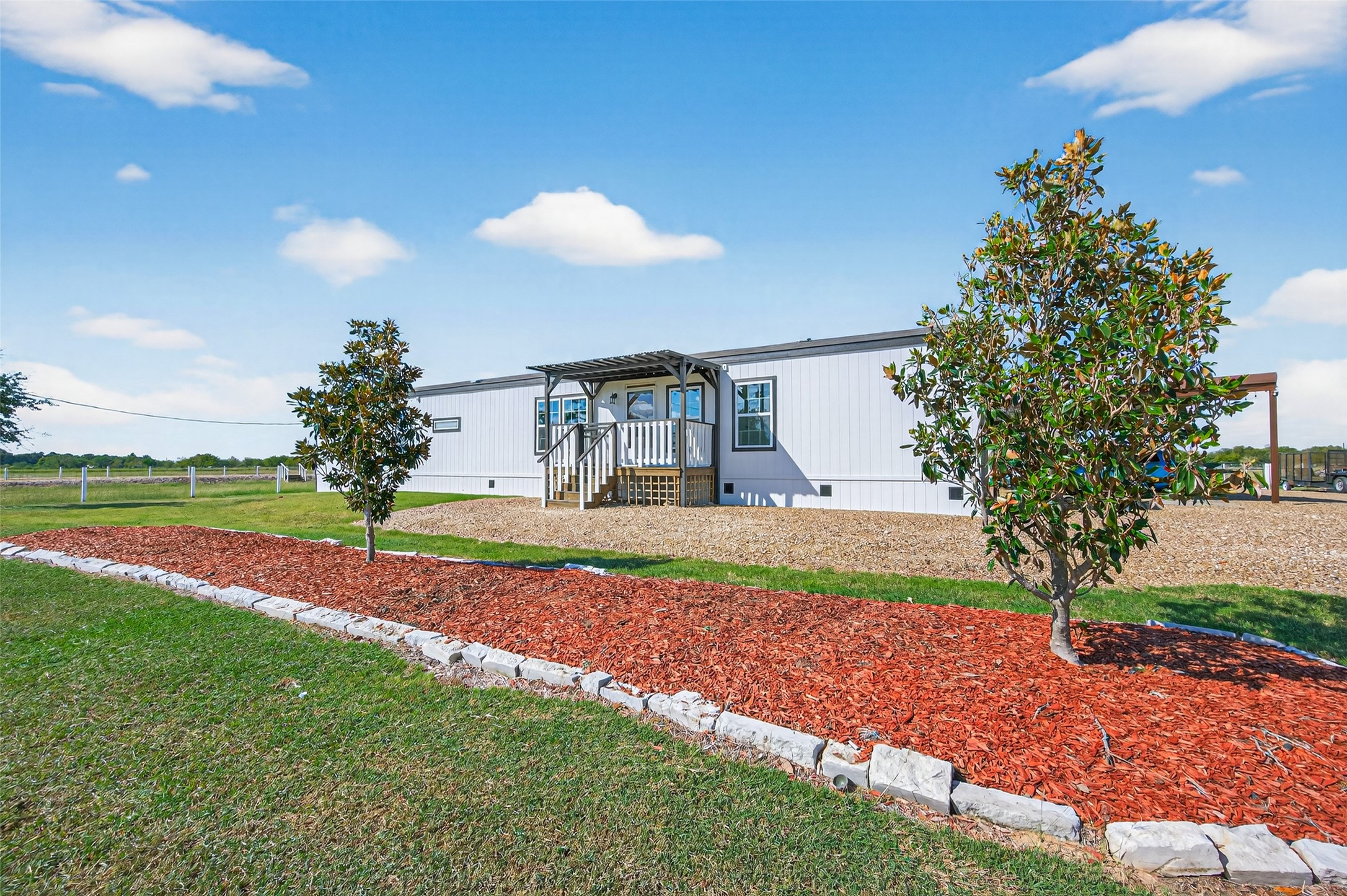 Front exterior view of the manufactured home set on open acreage, featuring a welcoming covered porch and wide-open rural surroundings.