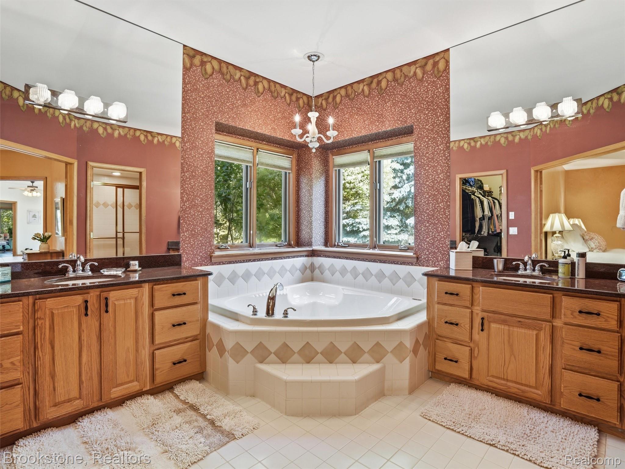 9523 Quail Ridge Run Brighton, MI 48114 - Photo 33 of 50 Corner soaking tub with tiled surround and multiple windows, flanked by dual wood vanities with dark countertops and integrated sinks