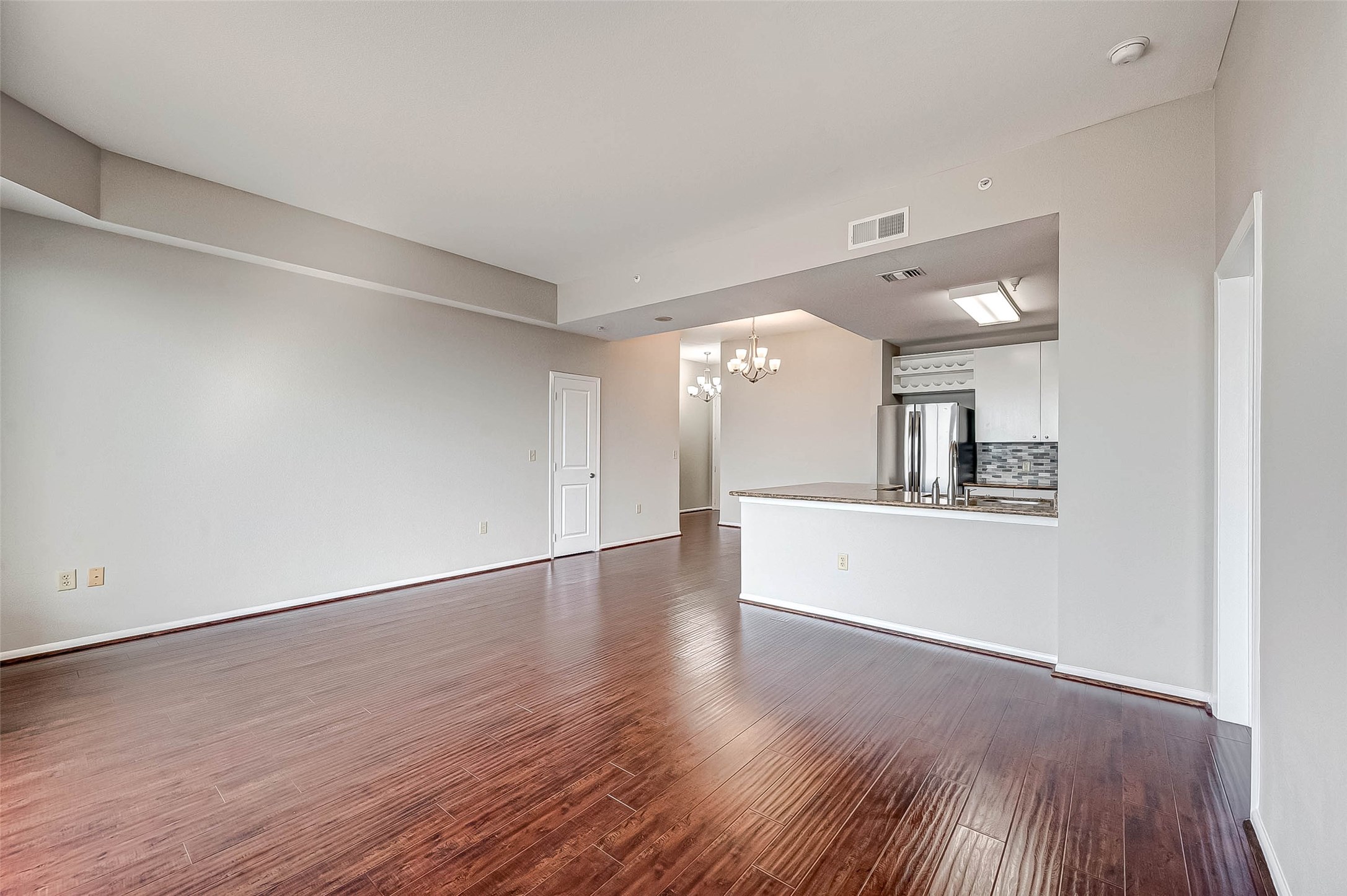 3505 Sage Road, Unit 2106 Houston, TX 77056 - Photo 8 of 31 This photo shows a spacious, open-concept living area with hardwood floors and neutral walls. It leads into a modern kitchen featuring stainless steel appliances, a tile backsplash, and a breakfast bar. A chandelier adds elegance, and there's ample natural light.