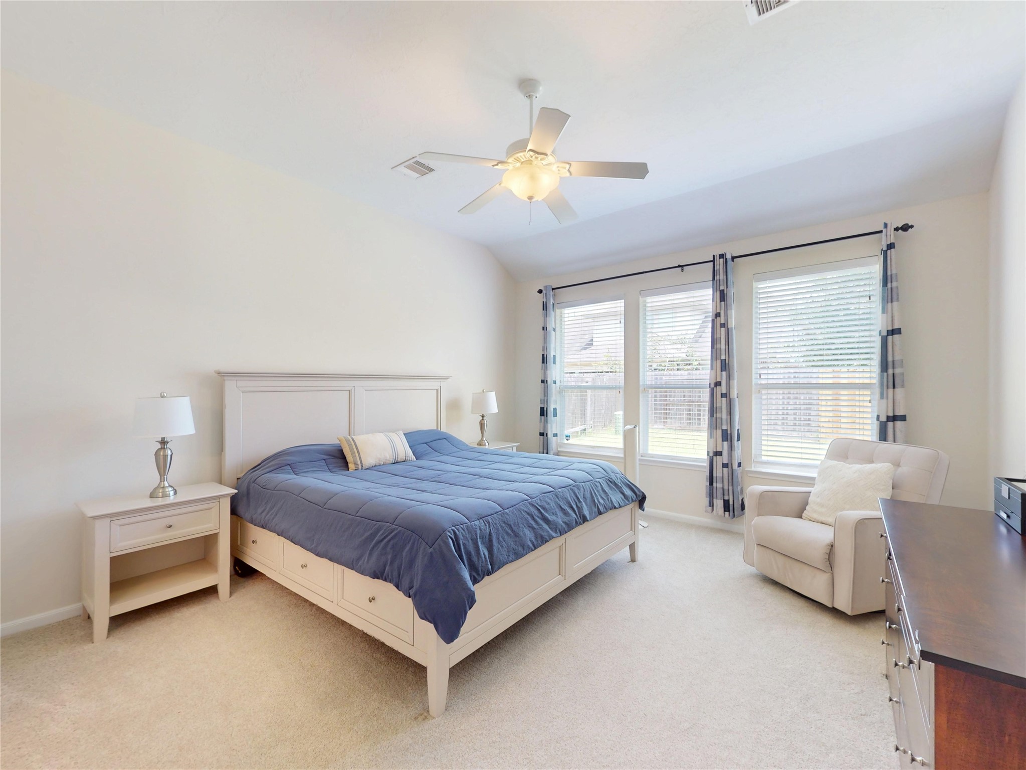17402 Rainer Valley Lane Humble, TX 77346 - Photo 15 of 33 This bright bedroom features a large bed with storage, two side tables with lamps, a cozy armchair, and a dresser. Natural light pours in through three windows adorned with striped curtains, and a ceiling fan adds comfort. The room is carpeted and painted in a neutral tone.