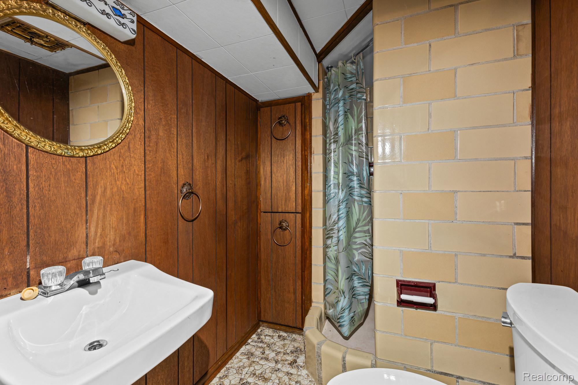 20261 Burt Road Detroit, MI 48219 - Photo 20 of 23 Bathroom featuring wood-finish paneling, a wall-mounted sink with a chrome faucet, a decorative mirror with an ornate frame, built-in cabinetry with ring pull hardware, and tiled shower walls