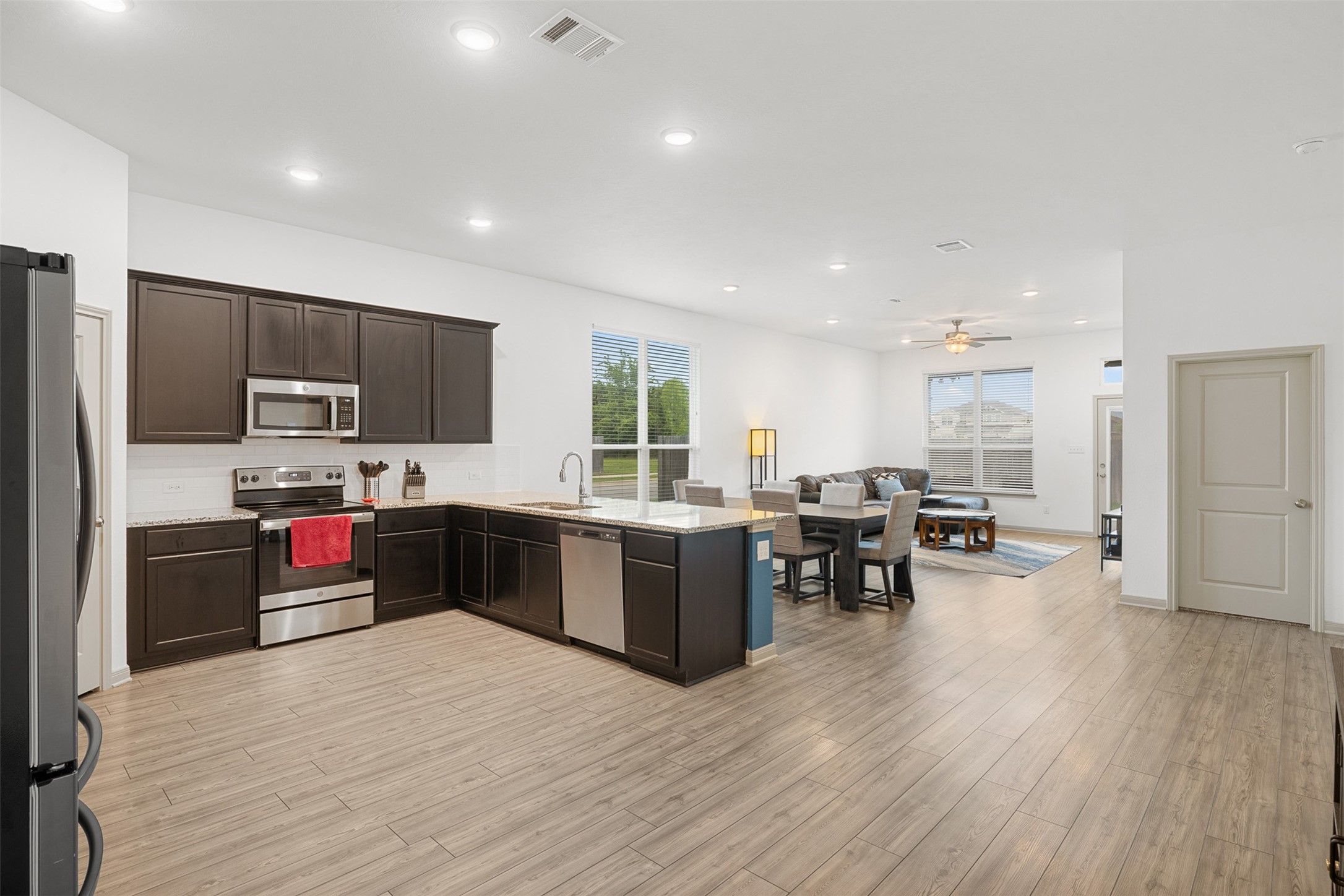 1968 Chief Street Bryan, TX 77807 - Photo 2 of 27 This photo showcases a modern open-concept living space with a sleek kitchen featuring dark cabinetry, stainless steel appliances, and an island. The area flows into a bright dining and living room with large windows, wood flooring, and recessed lighting, creating an inviting atmosphere.