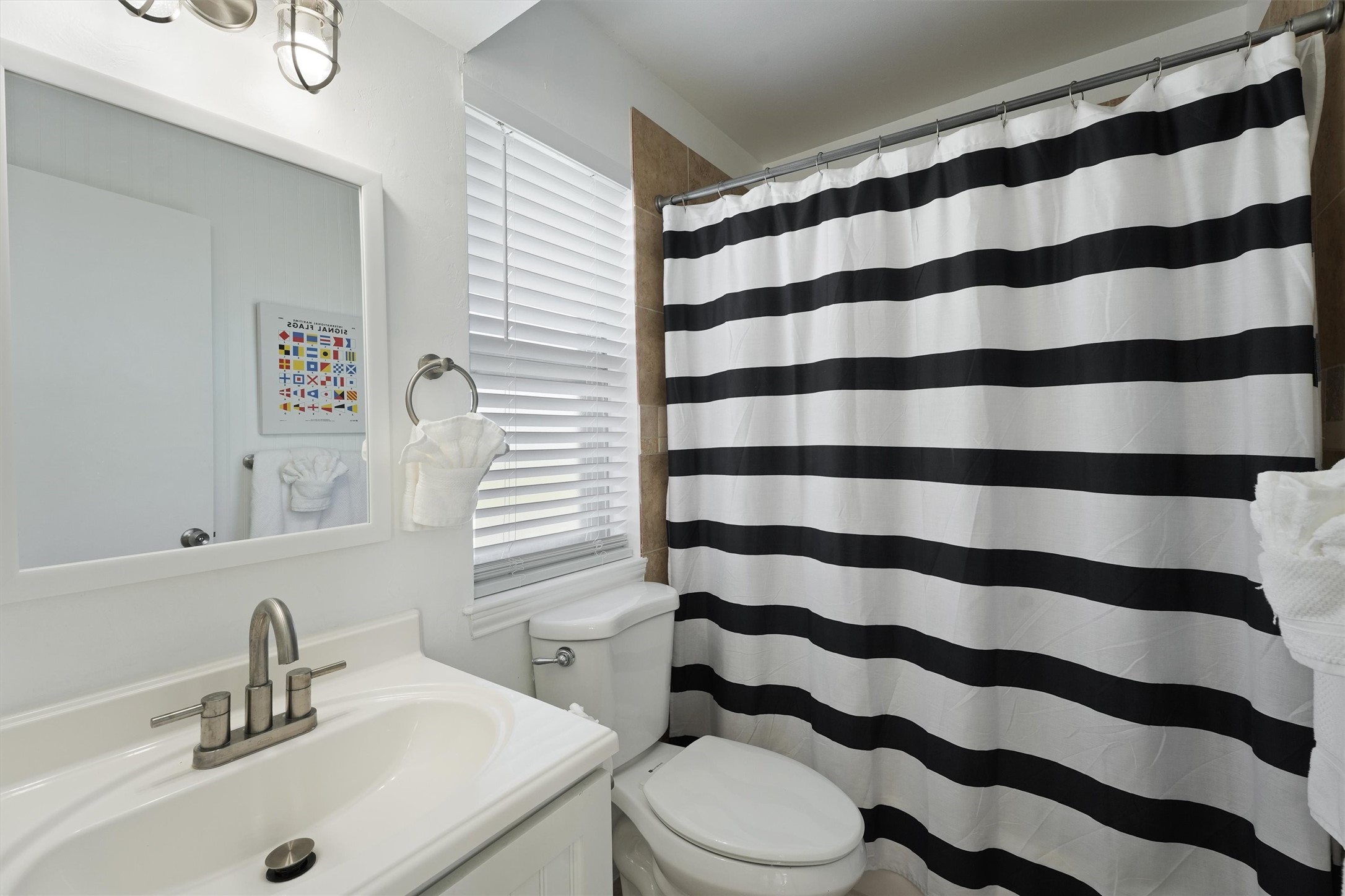 17607 Termini-San Luis Pass Road Galveston, TX 77554 - Photo 19 of 42 This full bathroom near the second floor living spaces features a modern vanity with a sleek brushed nickel faucet and a stylish framed mirror. The black-and-white striped shower curtain adds a bold, nautical touch, while natural light streams in through the window for a bright and inviting feel.